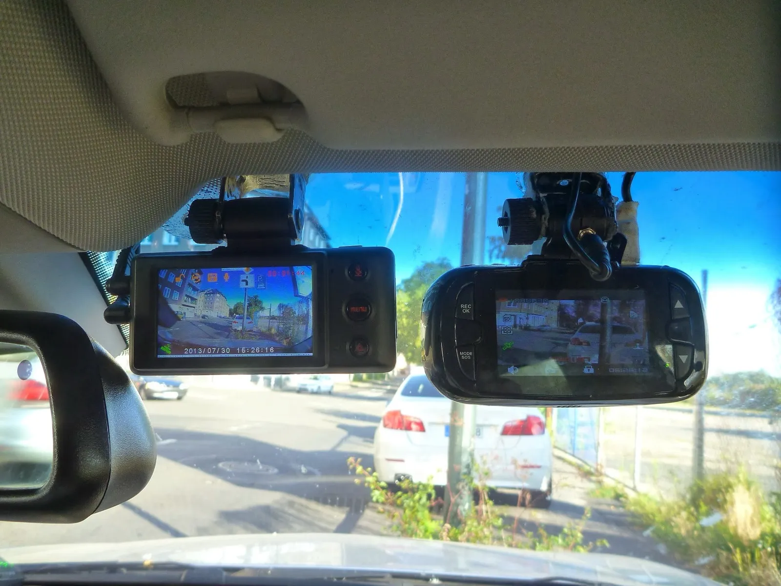 Two dash cameras mounted side by side at the top of a car windshield, their screens showing the street ahead