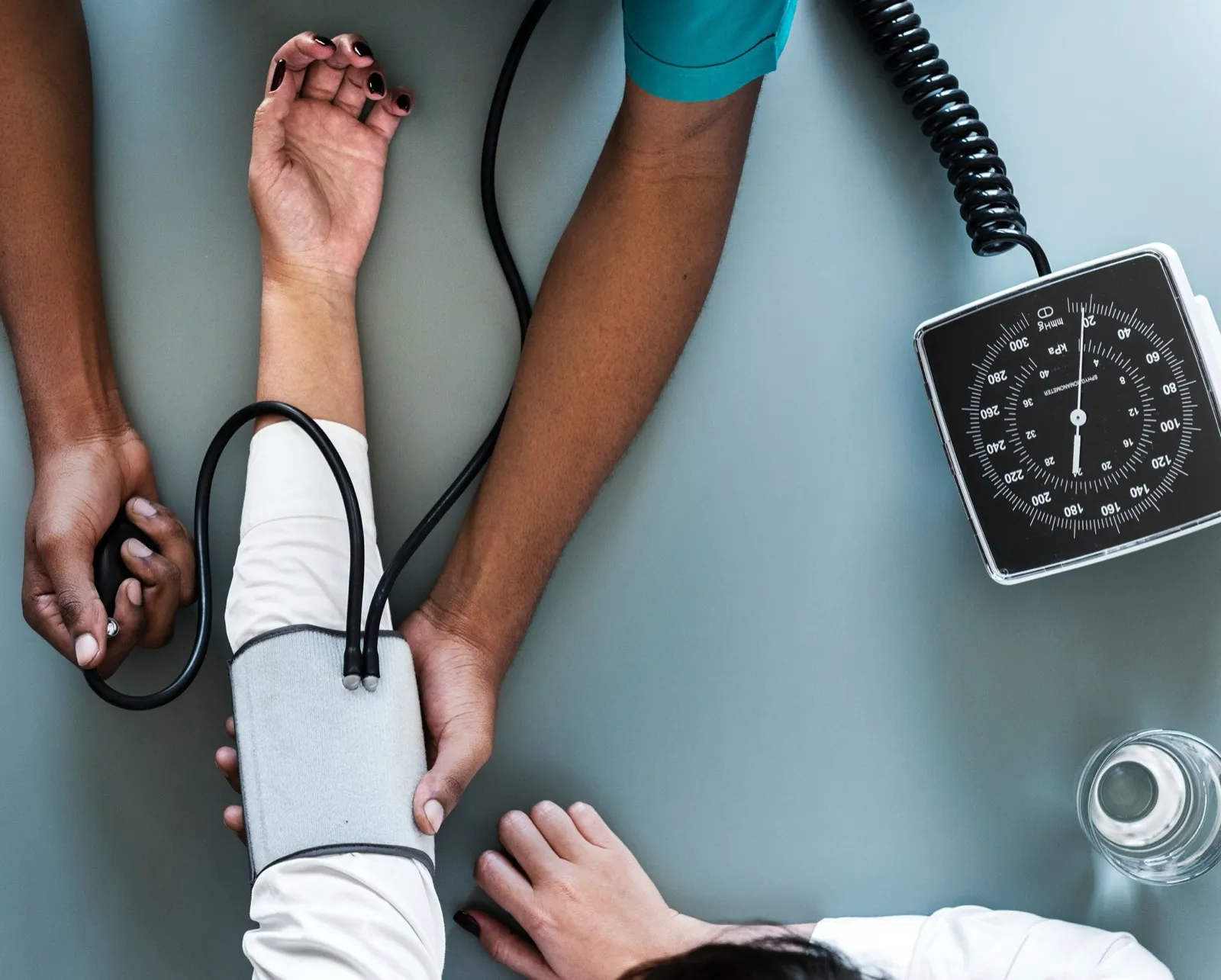 Seen from above, a clinician wraps a blood pressure cuff around a seated person's arm on a table beside a dial gauge