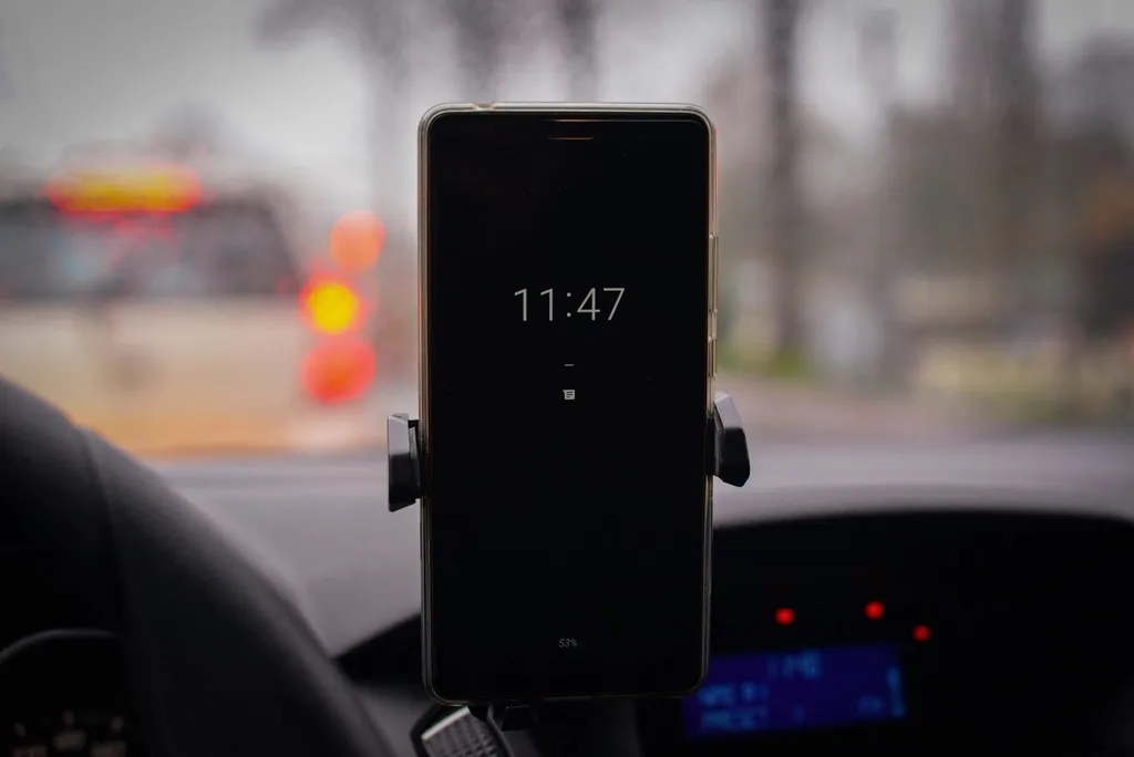 A smartphone held in a dashboard mount in front of the windshield, its screen showing the time, with traffic blurred ahead