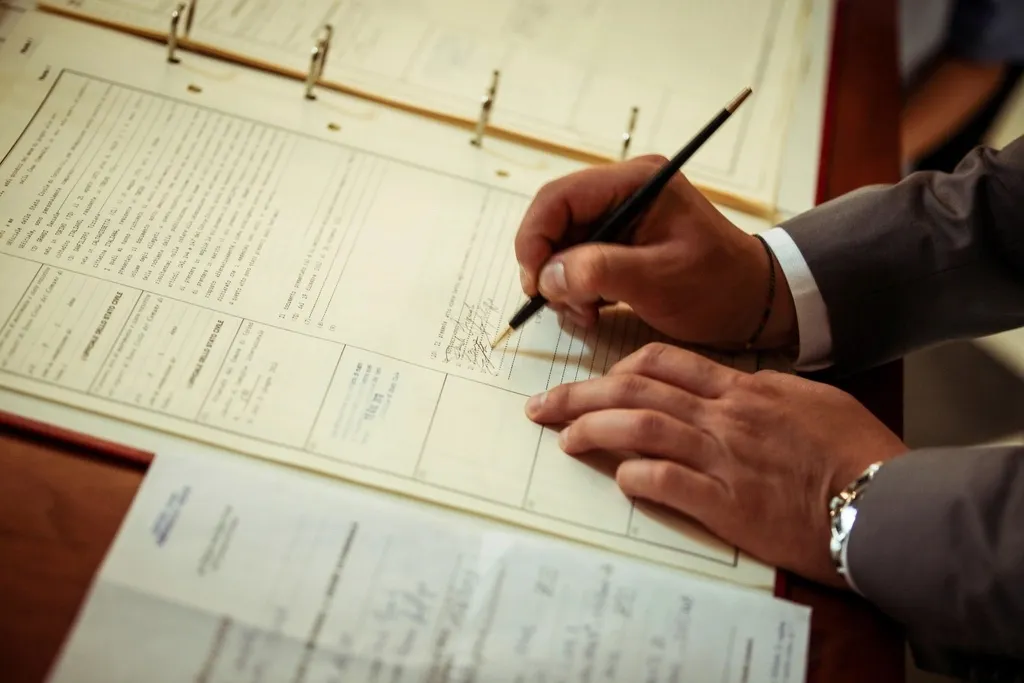 A man in a gray suit signs a page in an open ring binder with a black pen, with more printed forms on the desk