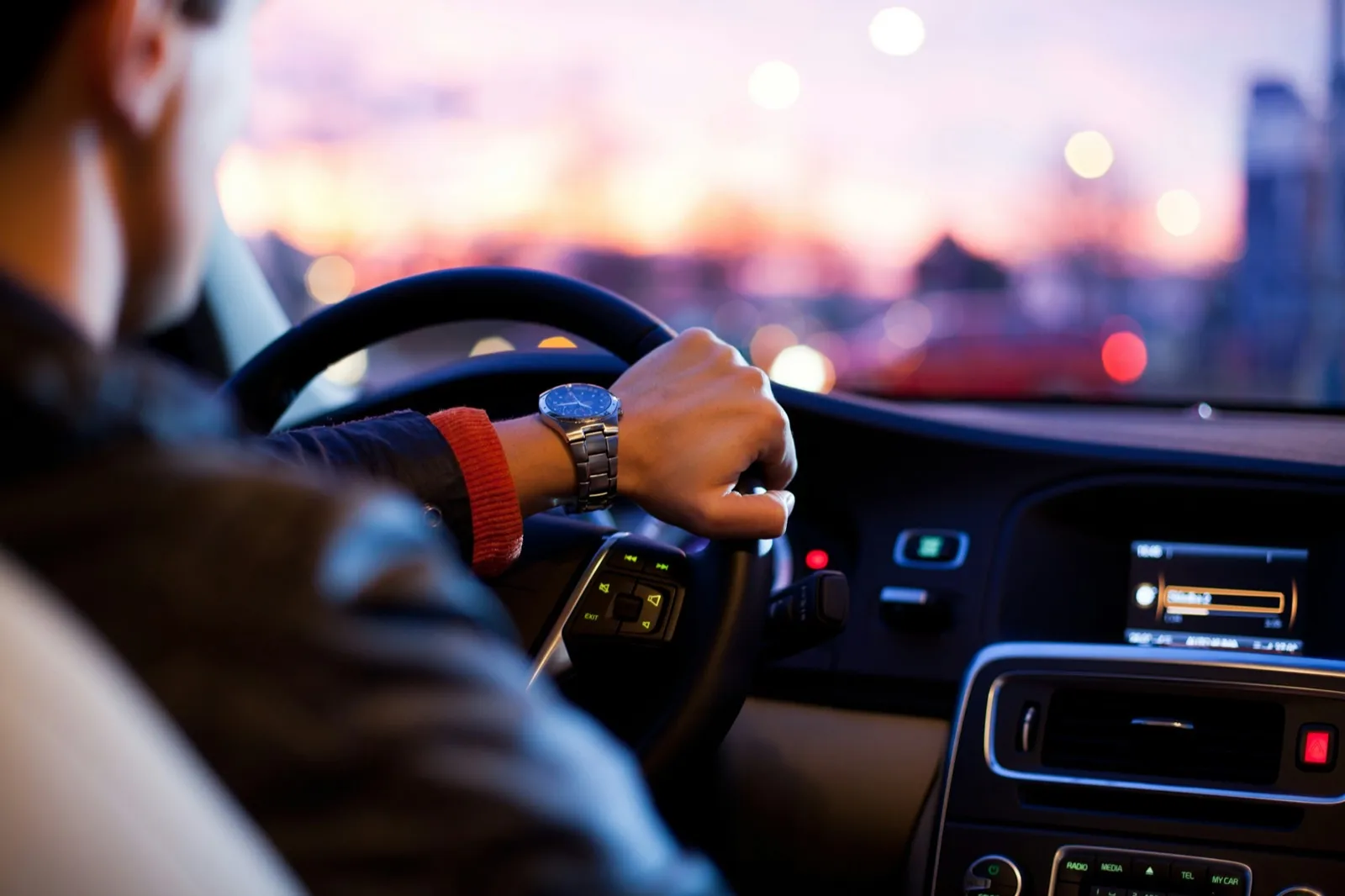 A driver wearing a wristwatch grips the steering wheel of a car at dusk, with city lights blurred through the windshield