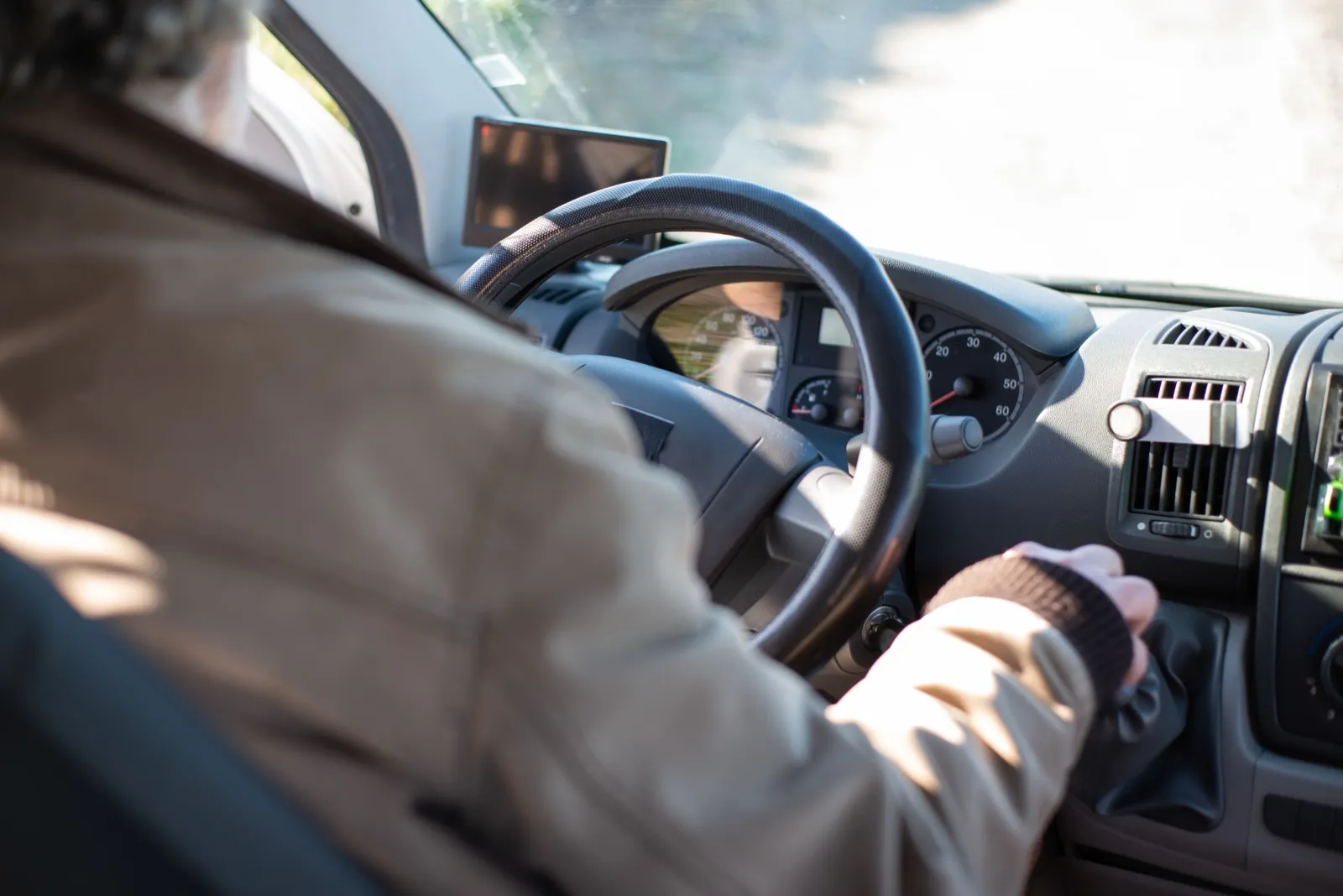 A gray-haired driver in a tan jacket, seen from behind the seat, reaching for the gear shift of a van on a sunny road