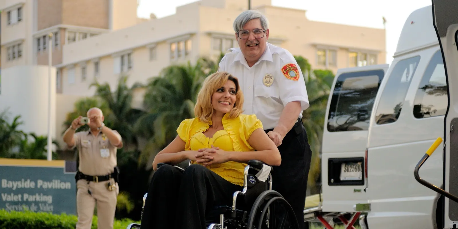 A smiling driver in a white uniform shirt pushes a woman in a wheelchair toward a white lift van outside a hospital