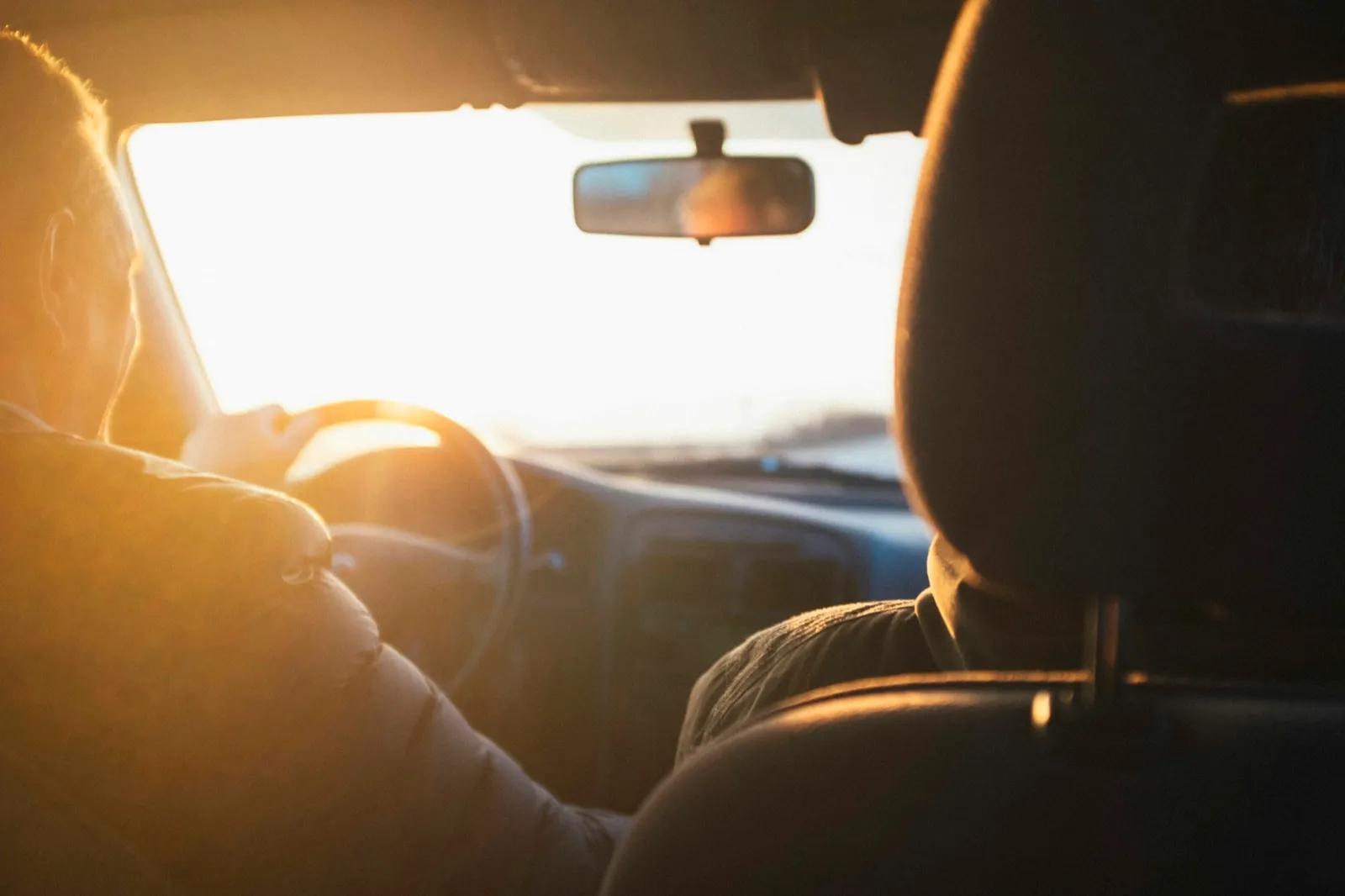 A driver seen from the back seat holding the steering wheel as low sun shines through the windshield