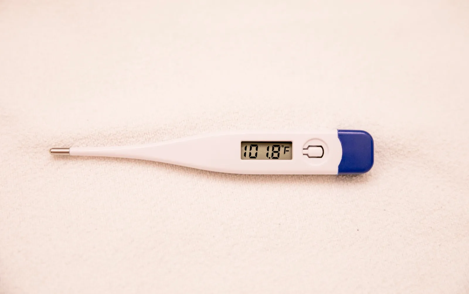 A white digital thermometer with a blue cap lying on a cream towel, its display reading a fever of 101.8 degrees