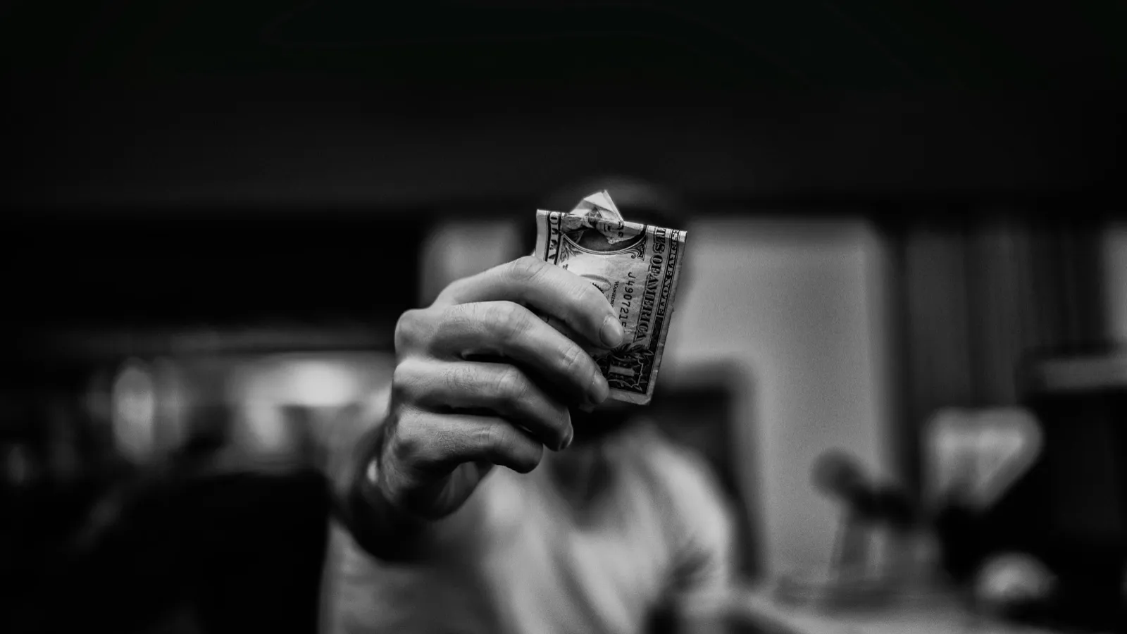 A hand holding out a folded one-dollar bill toward the camera, in black and white