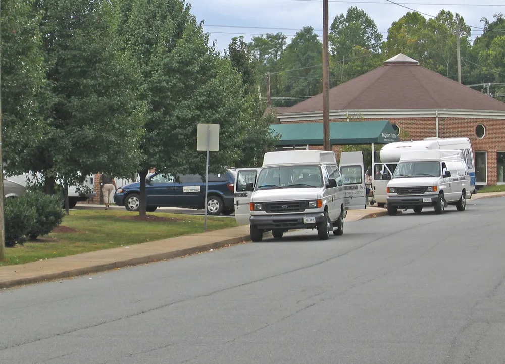 White passenger vans parked along a curb outside a brick building, with their side doors open for riders
