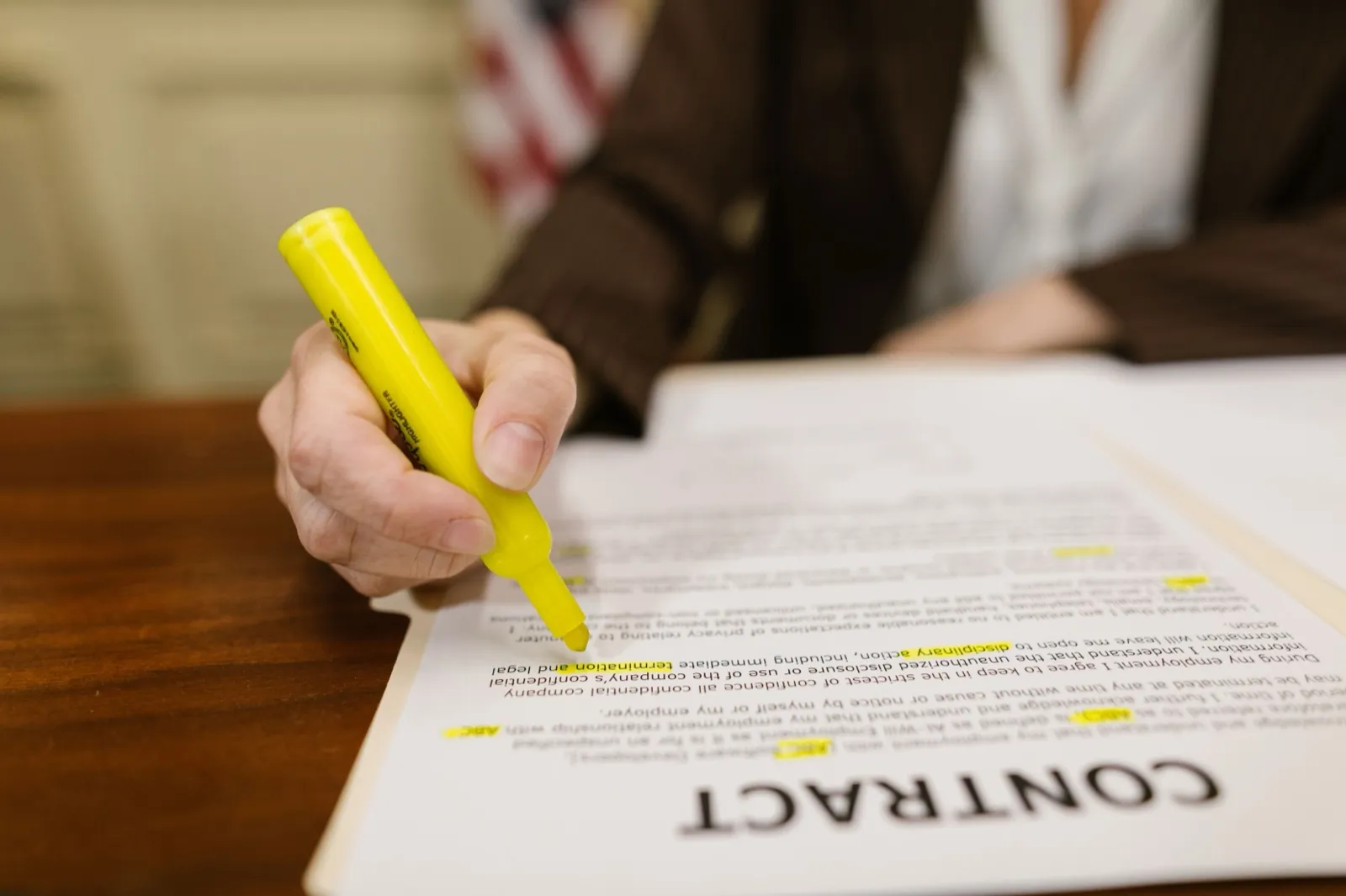 A hand marks terms in a printed contract with a yellow highlighter on a wooden desk