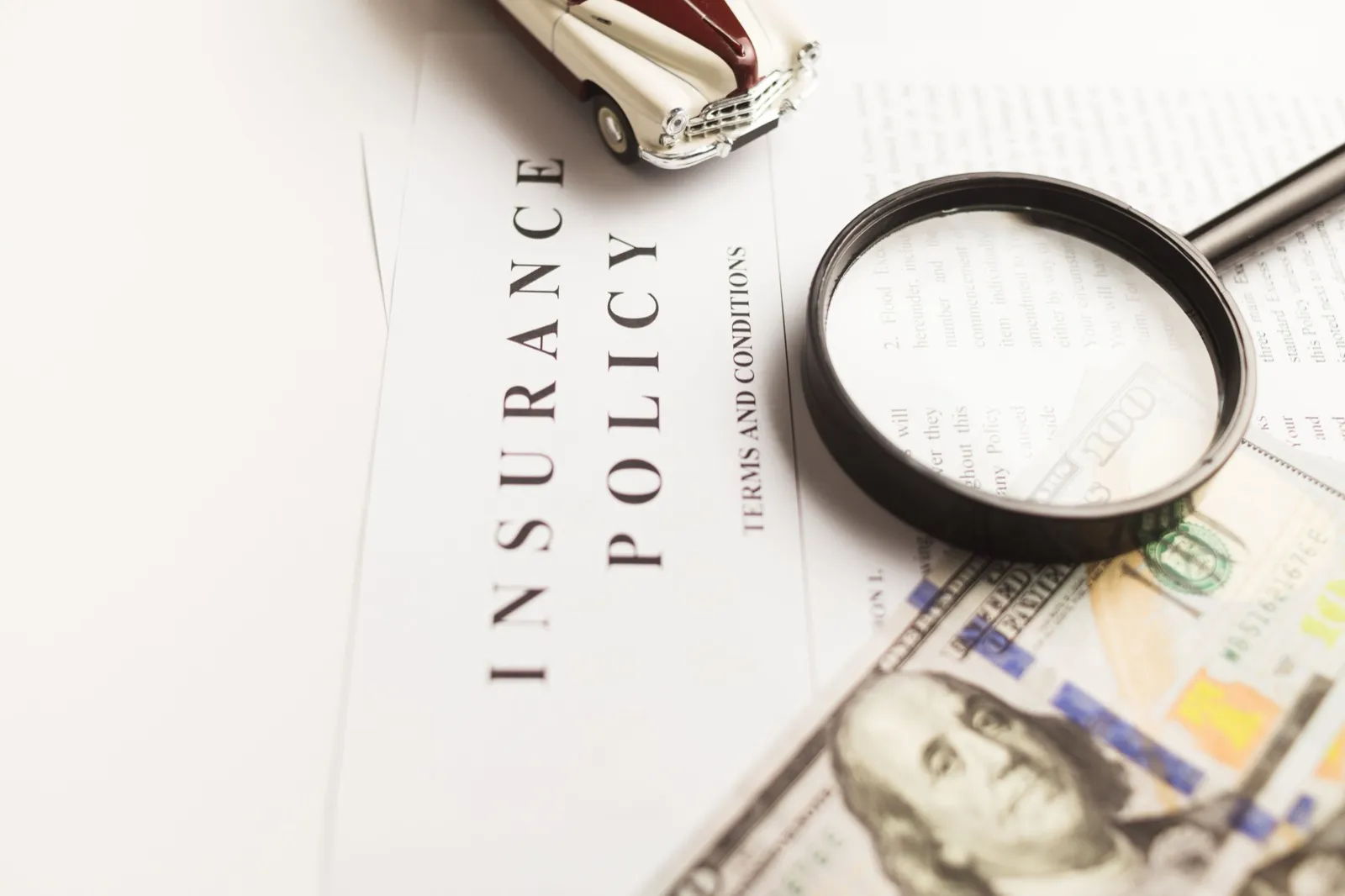 An insurance policy document on a white desk with a magnifying glass, a hundred-dollar bill, and a toy car