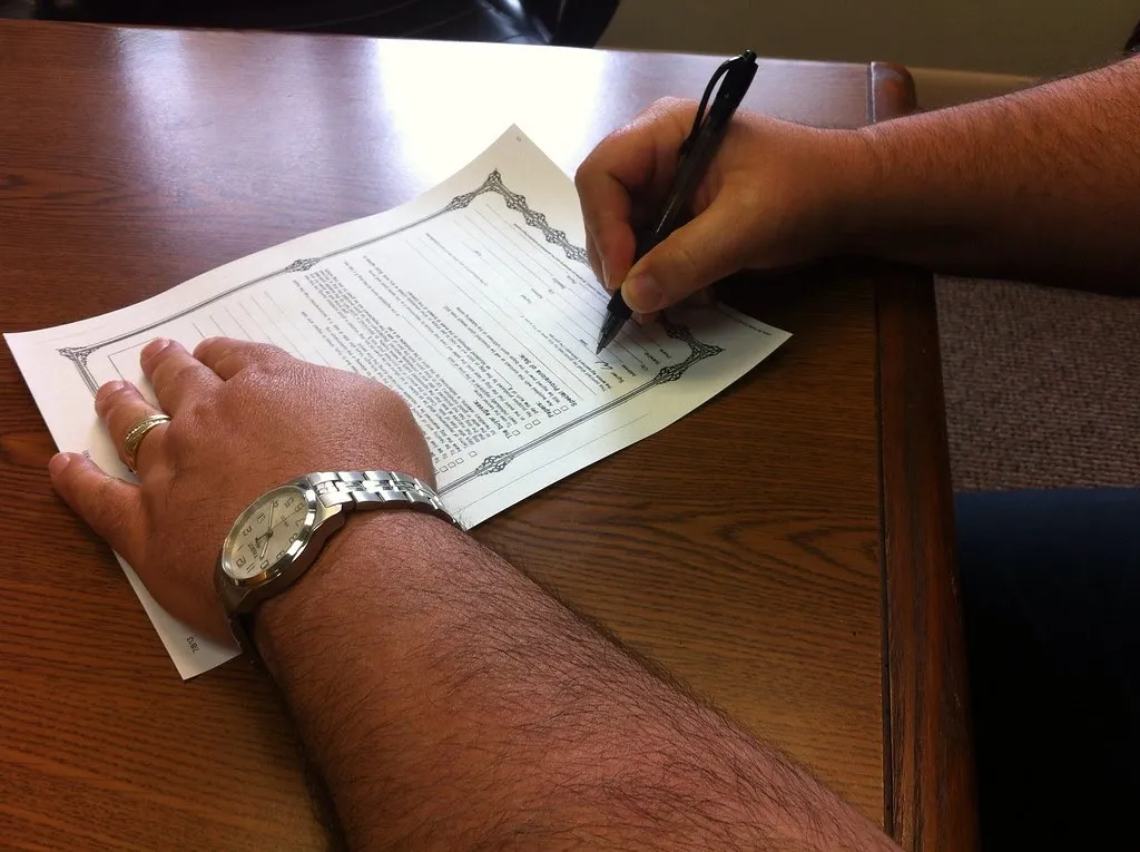 A person wearing a wristwatch fills out a printed form with a pen at a wooden desk