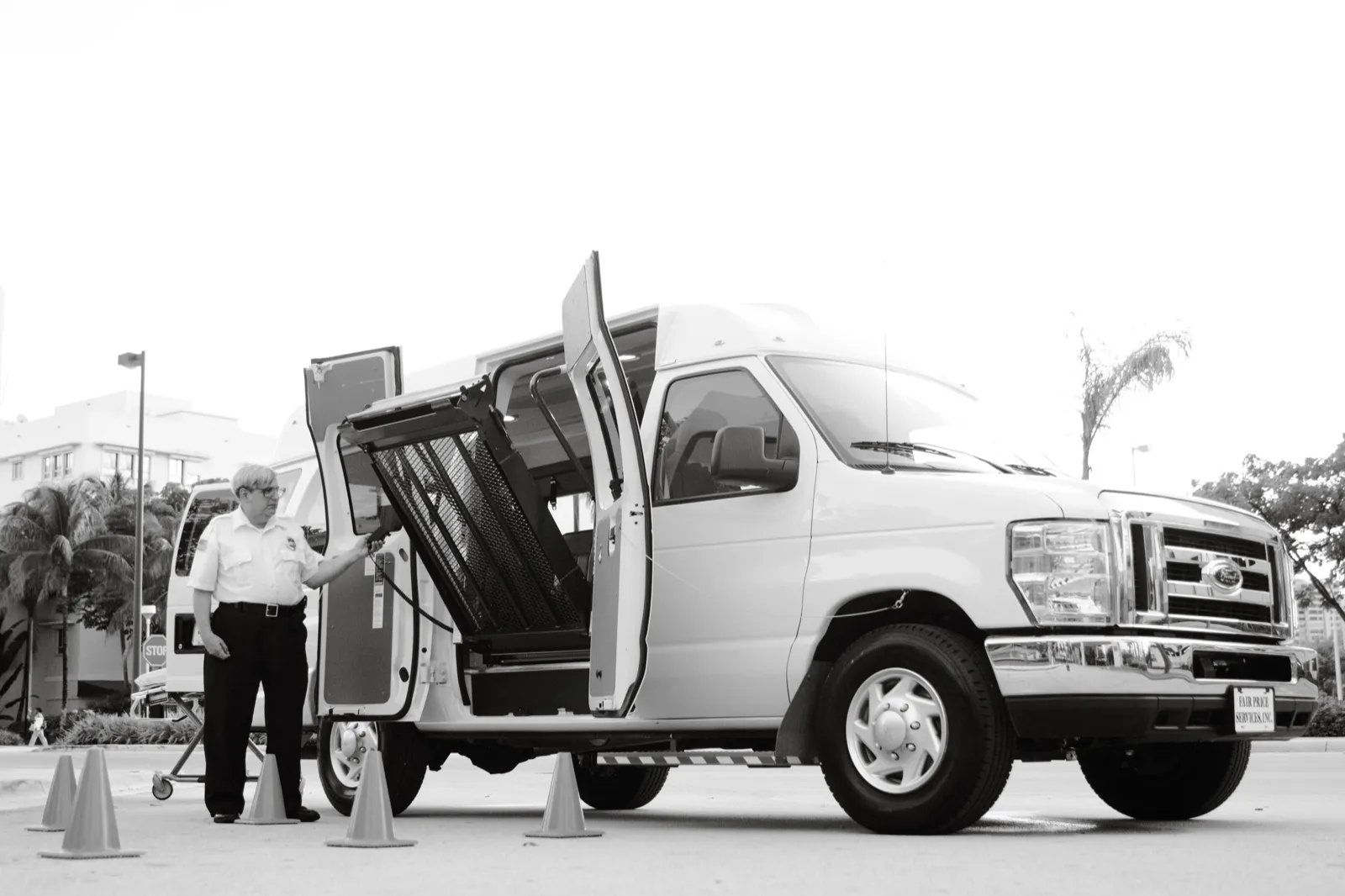 Black-and-white photo of a driver in a uniform shirt using a hand control to work the wheelchair lift of a white van, with safety cones set around it