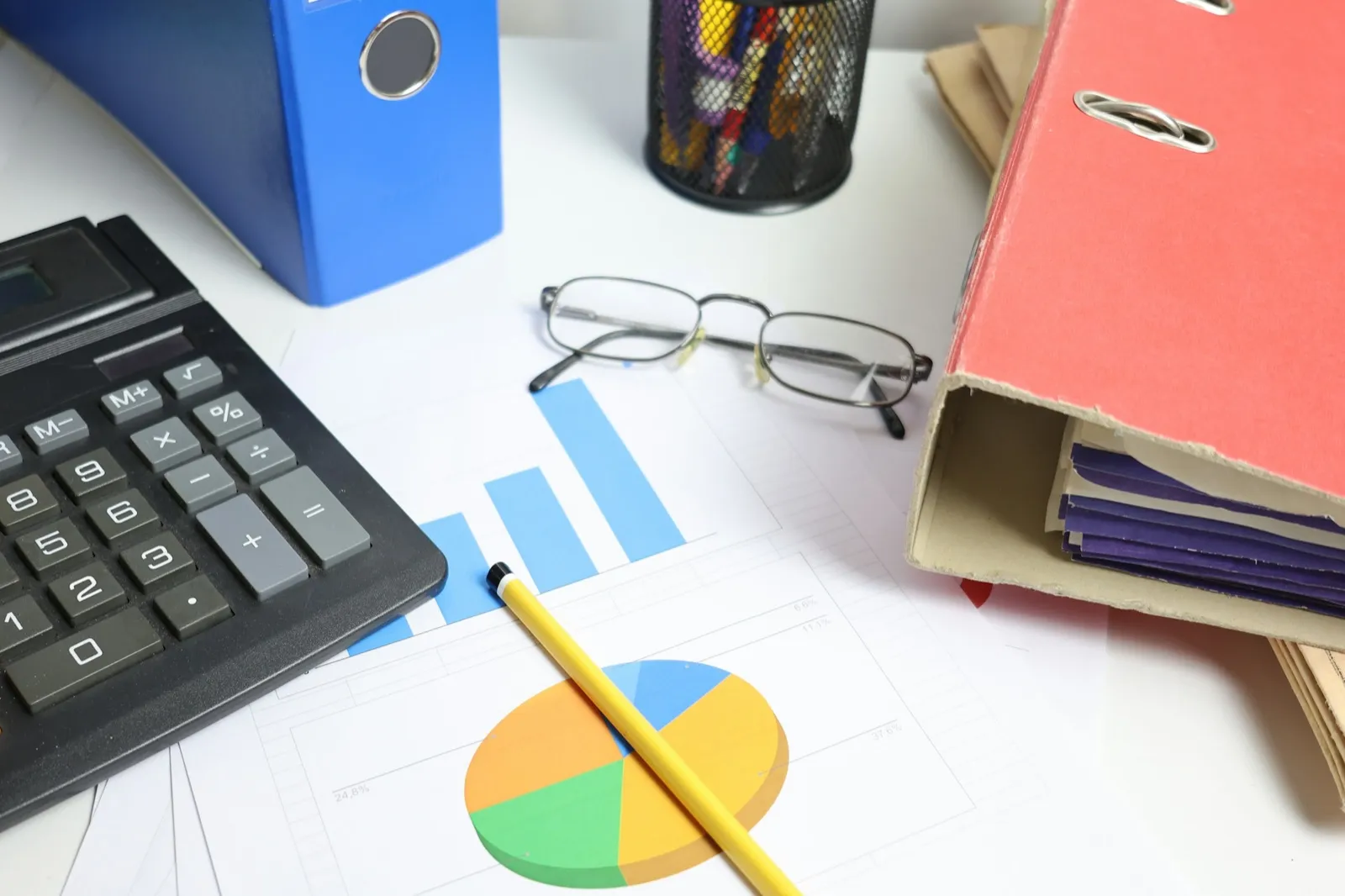 Printed bar and pie charts on a desk beside a calculator, reading glasses, a pencil, and two office binders