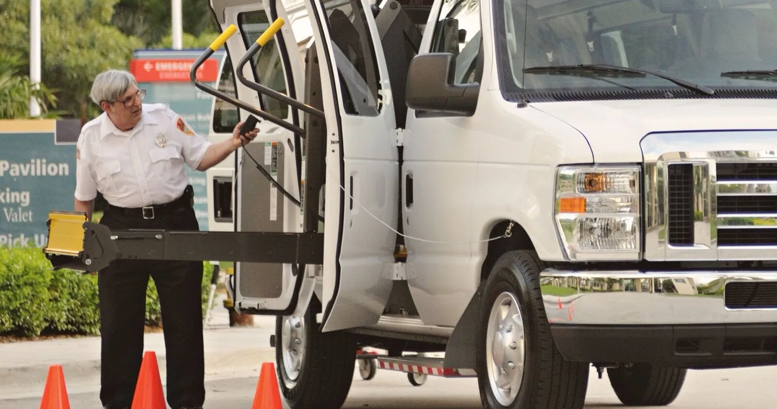 A driver in a white uniform shirt works the controls of a wheelchair lift at the open side doors of a white van, with orange cones set on the pavement