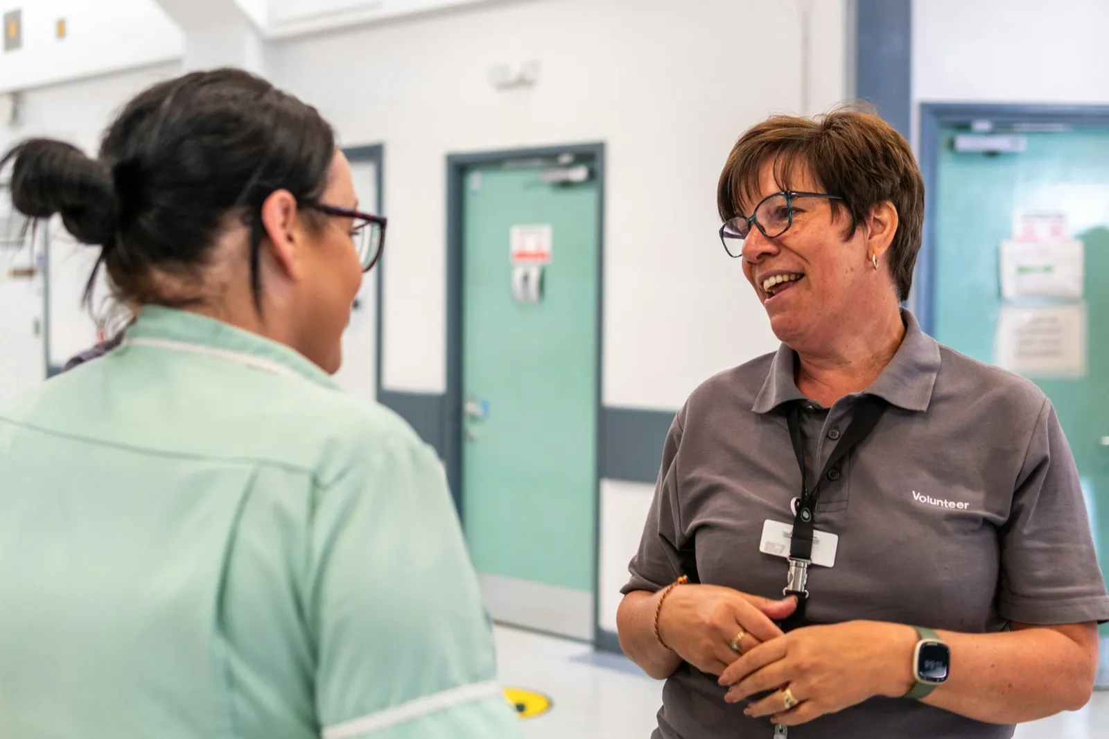 A smiling woman in a gray polo shirt with a lanyard talks with a nurse in a pale green uniform in a hospital hallway
