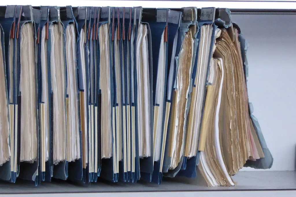 A row of worn blue hanging file folders stuffed with aging paper records on a shelf