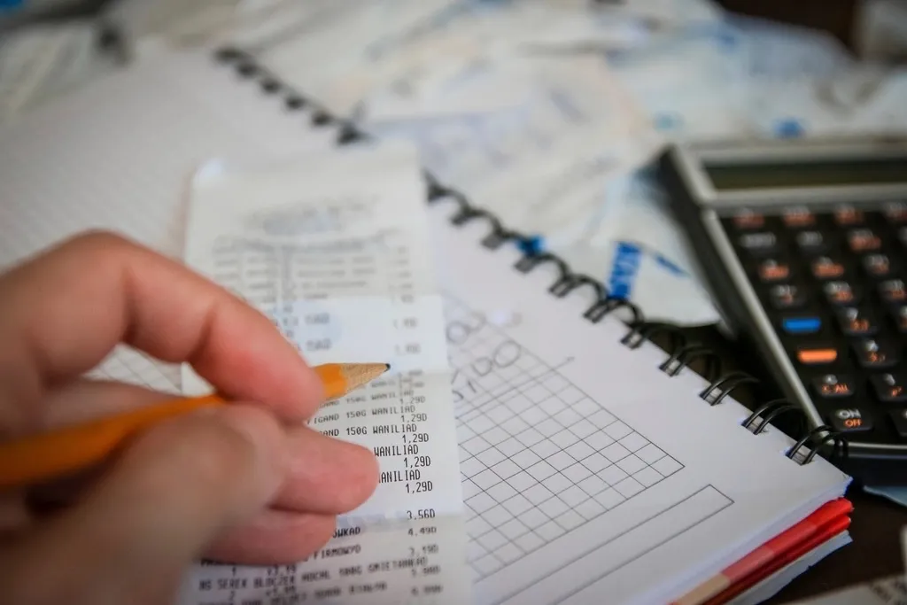 A hand holding a pencil over a printed receipt, next to a notebook and a desk calculator