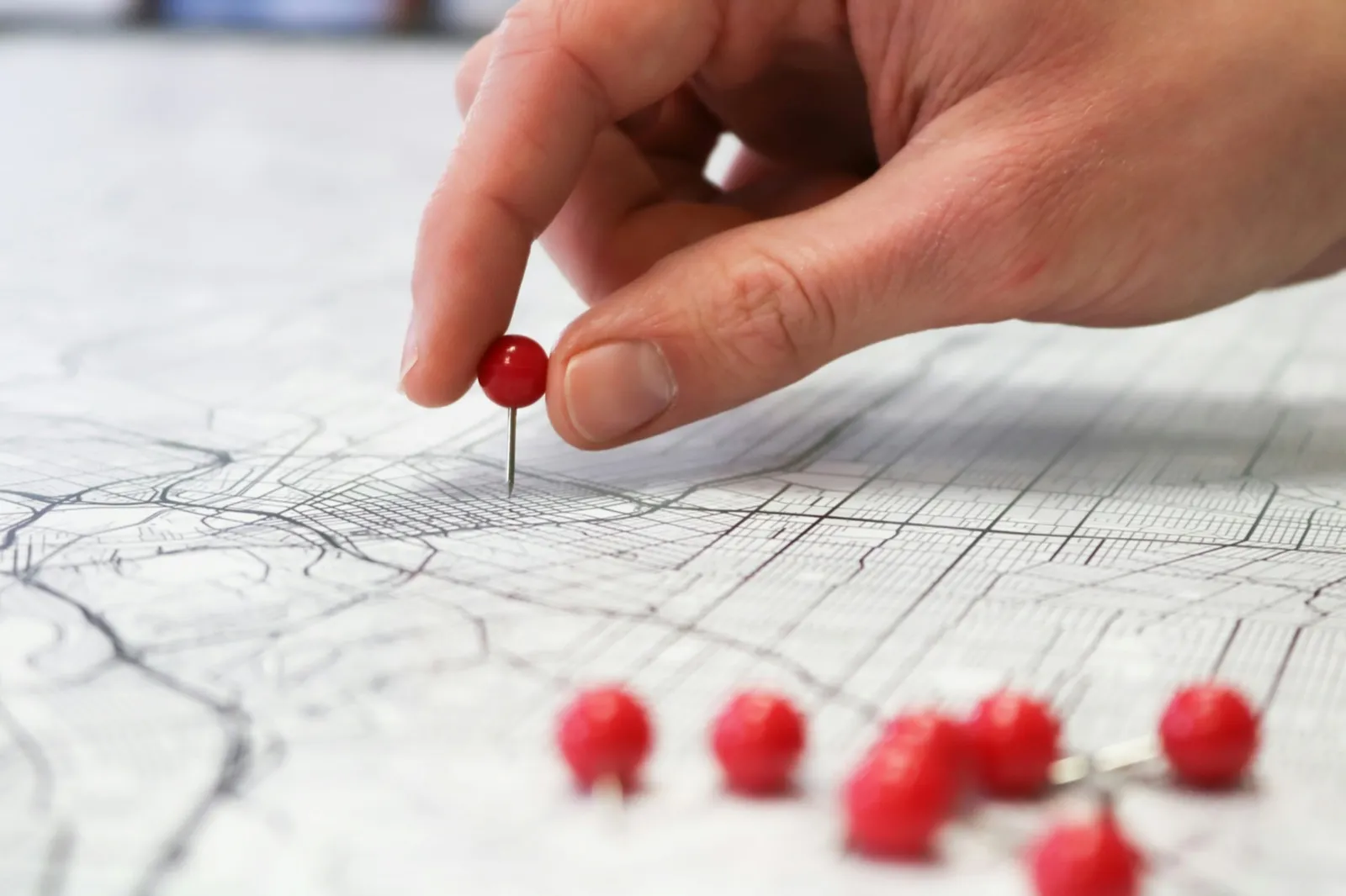 A hand pressing a red pin into a paper street map of a city, with more red pins lying on the map