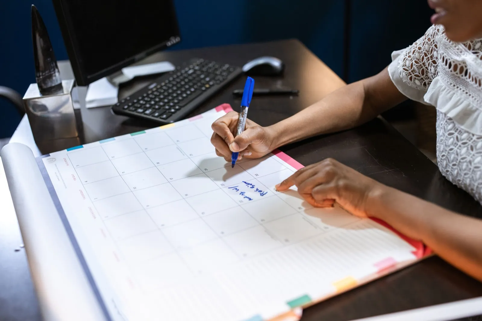 A woman at an office desk writing with a marker on a large paper desk calendar, with a computer keyboard behind it