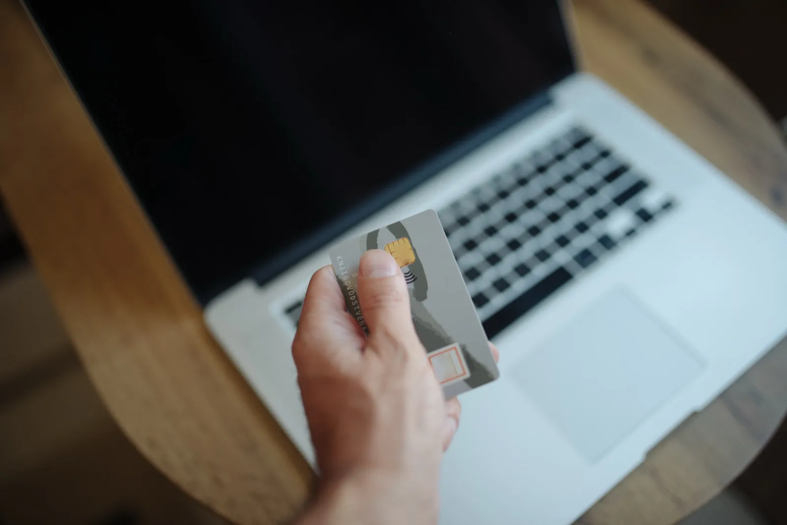A hand holding a payment card in front of an open laptop on a wooden table