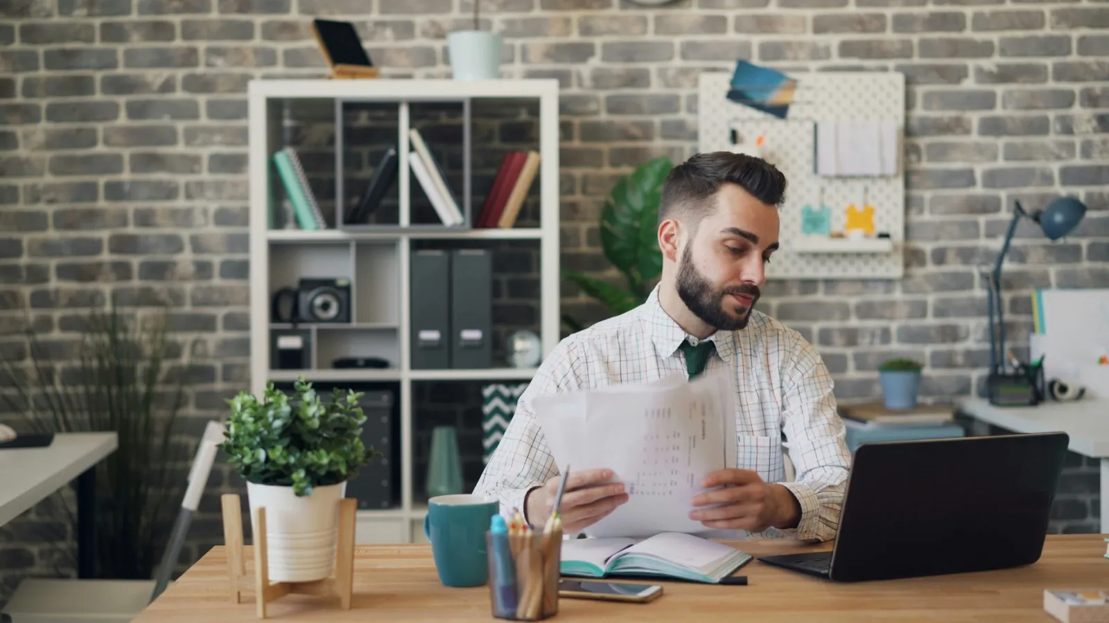 A man at an office desk reads printed pages with a table on them while working on an open laptop