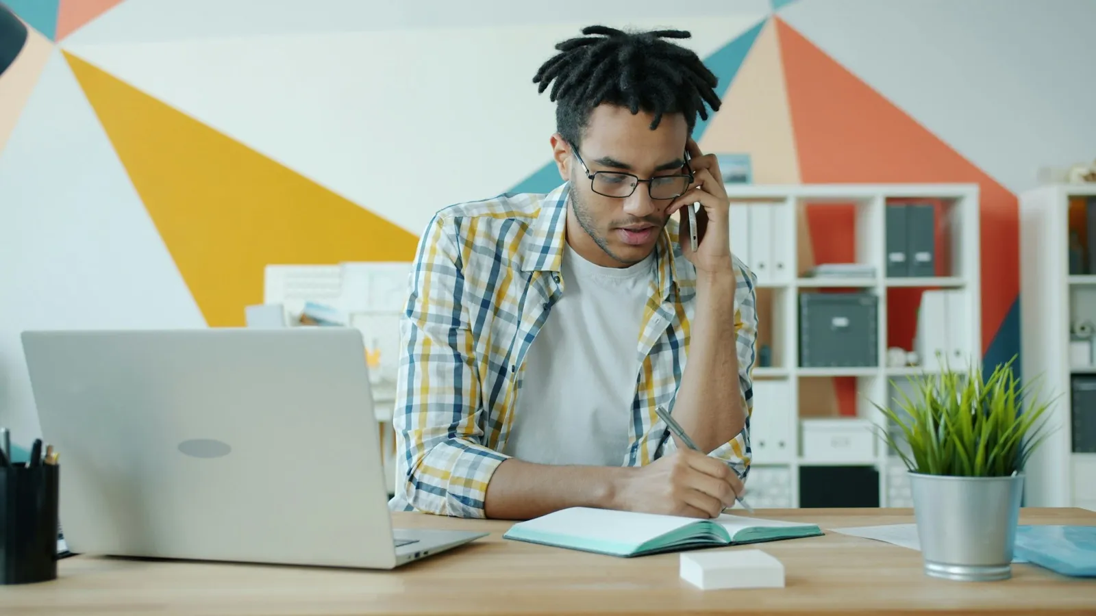 A man at an office desk talking on a phone and writing notes in a notebook, with a laptop beside him
