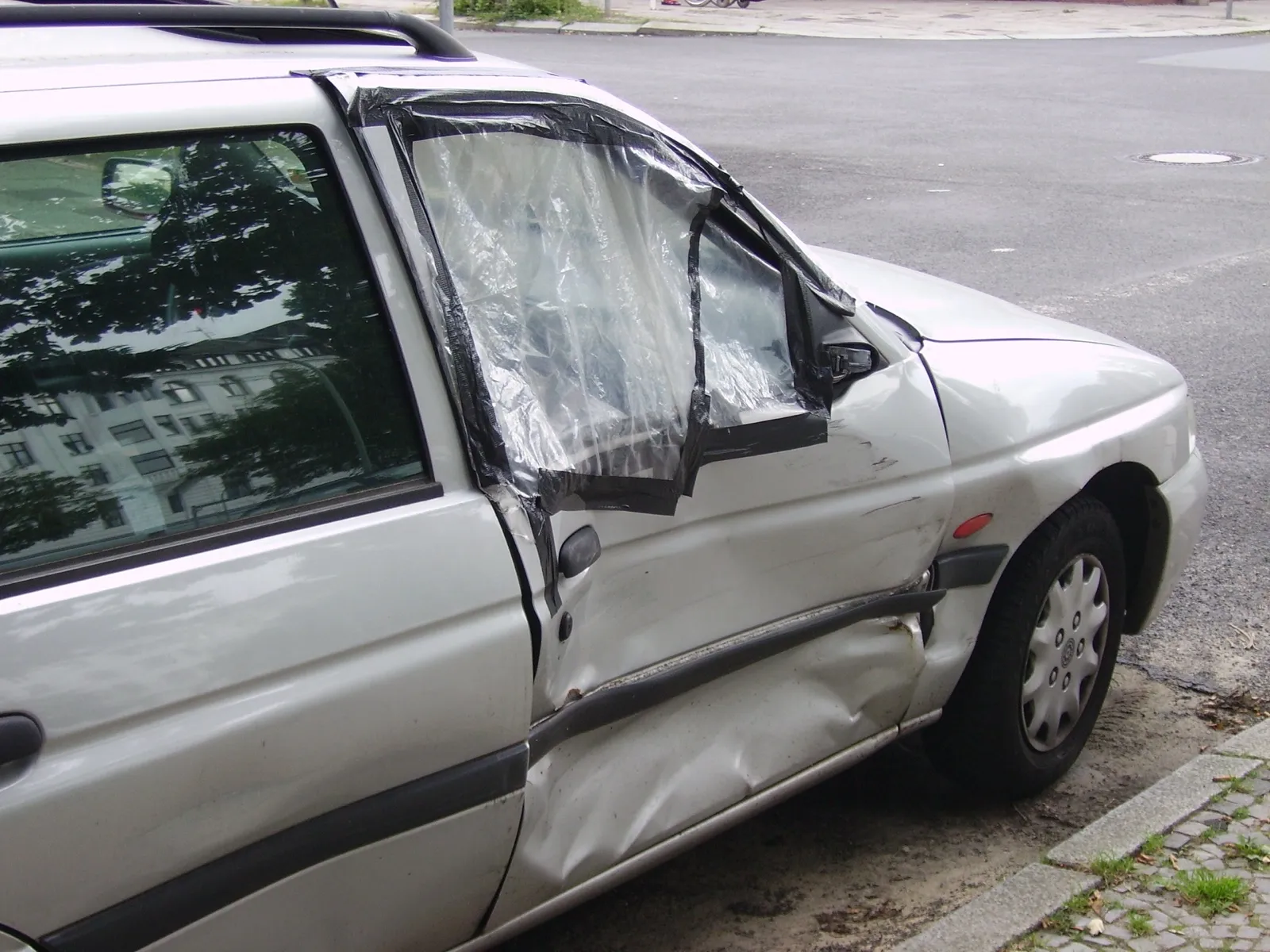 A silver car parked at a curb with a dented front passenger door and its broken window covered in plastic and tape