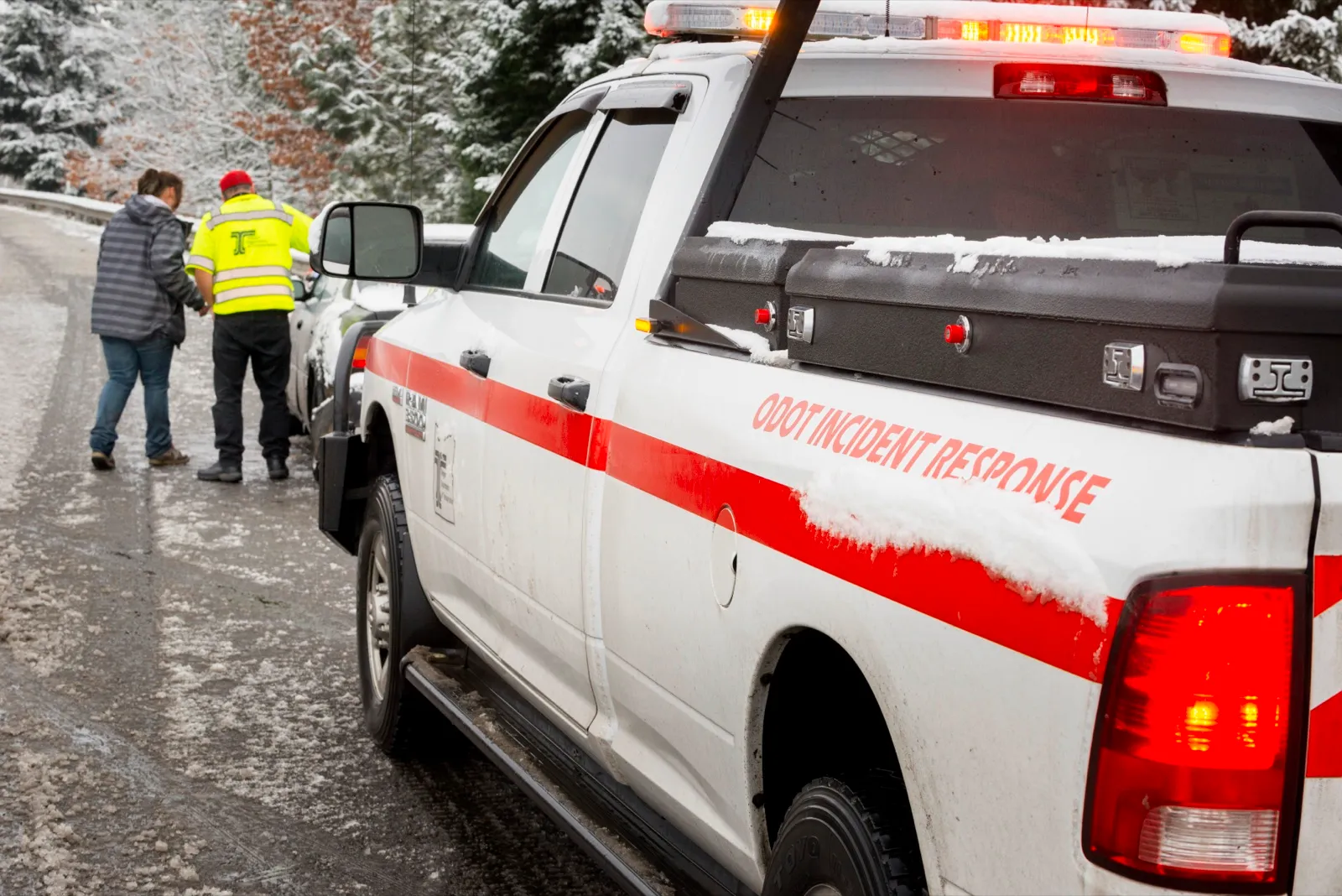 A state highway incident response truck with its lights on, stopped behind a car on a snowy roadside while a responder talks with the driver