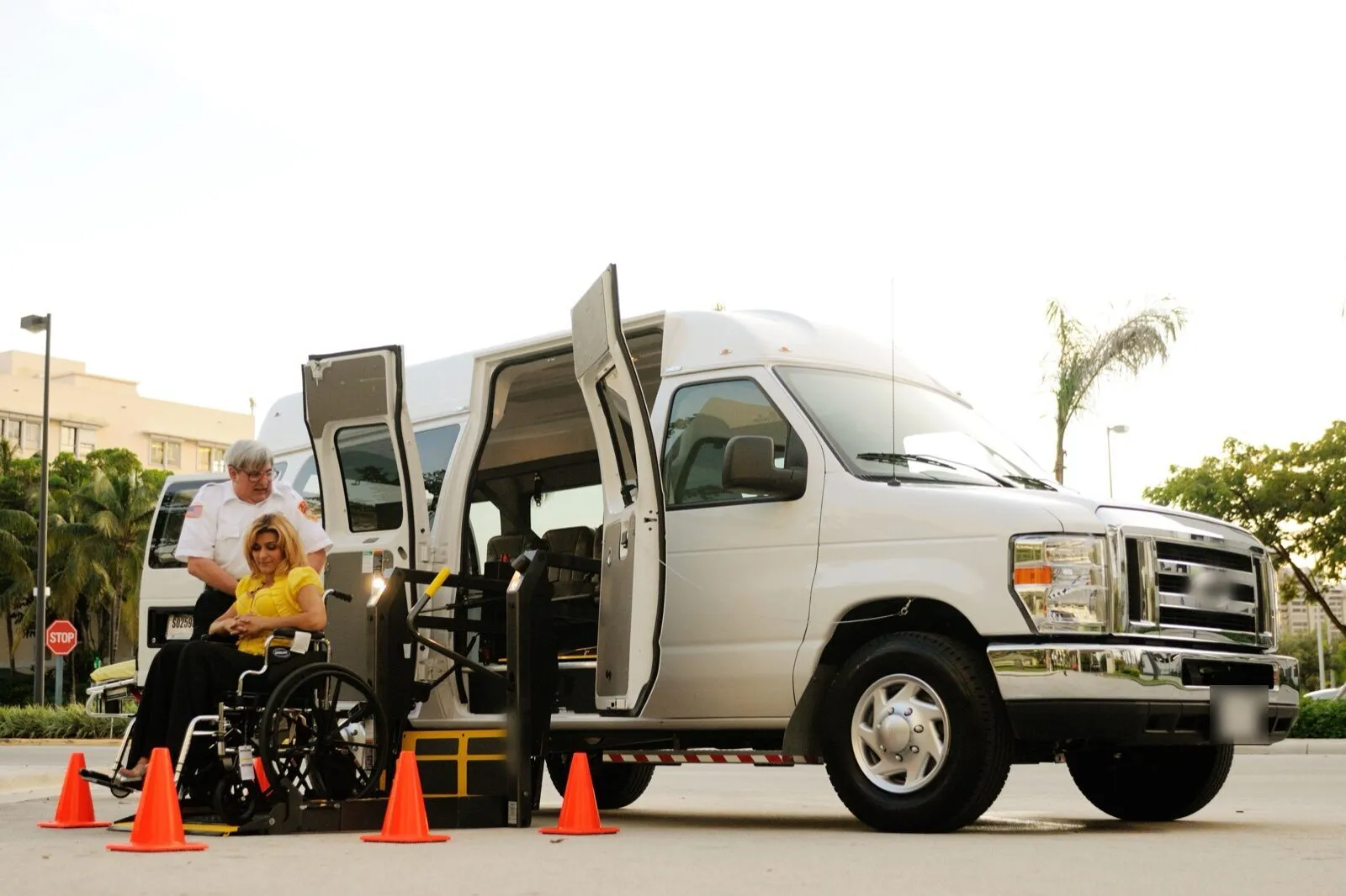 A woman in a wheelchair on the lift of a white high-roof van, with a uniformed driver beside her and orange cones around the lift