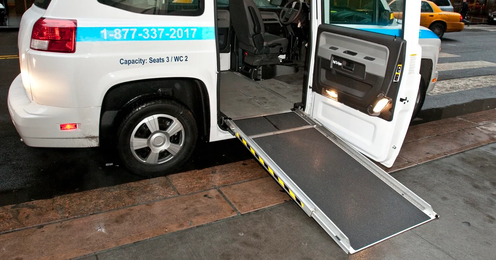 A white wheelchair van with a phone number and its seating capacity lettered on the side, its side door open and a ramp lowered to the curb