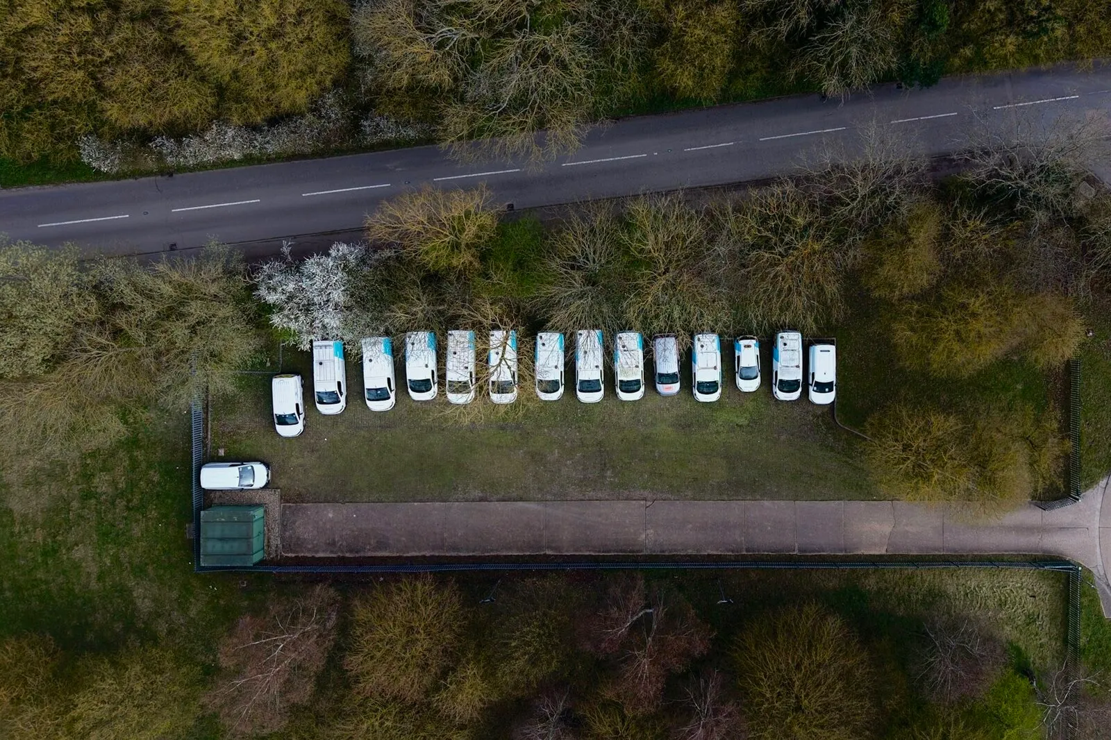 An aerial view of a row of white vans parked side by side in a grassy lot beside a two-lane road
