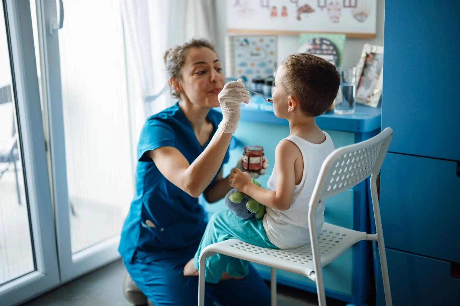 A health worker in blue scrubs kneels beside a young boy on a chair in a pediatric clinic and gives him a spoonful of medicine