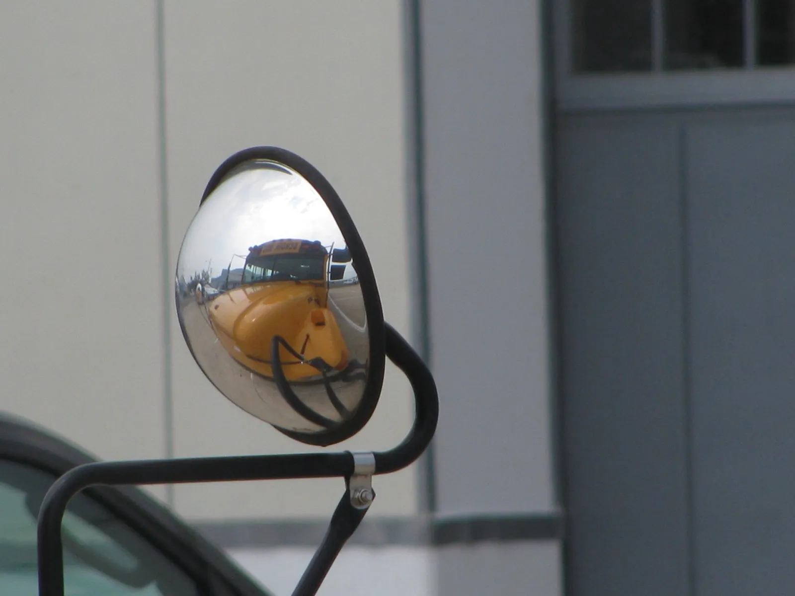 A round chrome crossview mirror on its bracket at the front of a yellow school bus, reflecting the bus hood