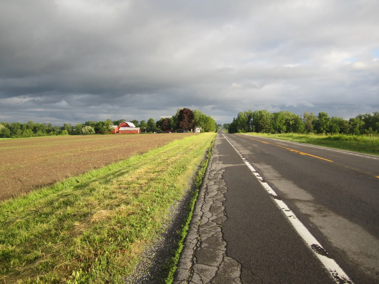 A two-lane rural highway in upstate New York running past a plowed field and a red barn under heavy clouds