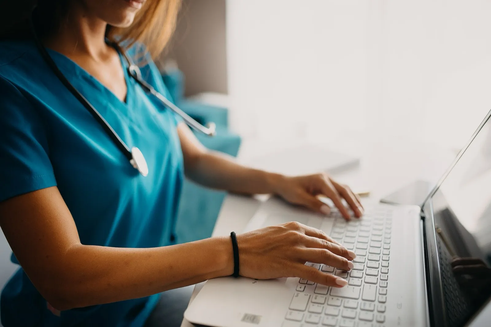 A health care worker in teal scrubs with a stethoscope around her neck typing on a white laptop at a desk