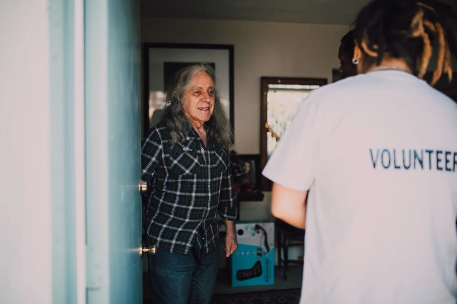 A volunteer in a white T-shirt printed with the word Volunteer stands at the open front door of an older man with long gray hair