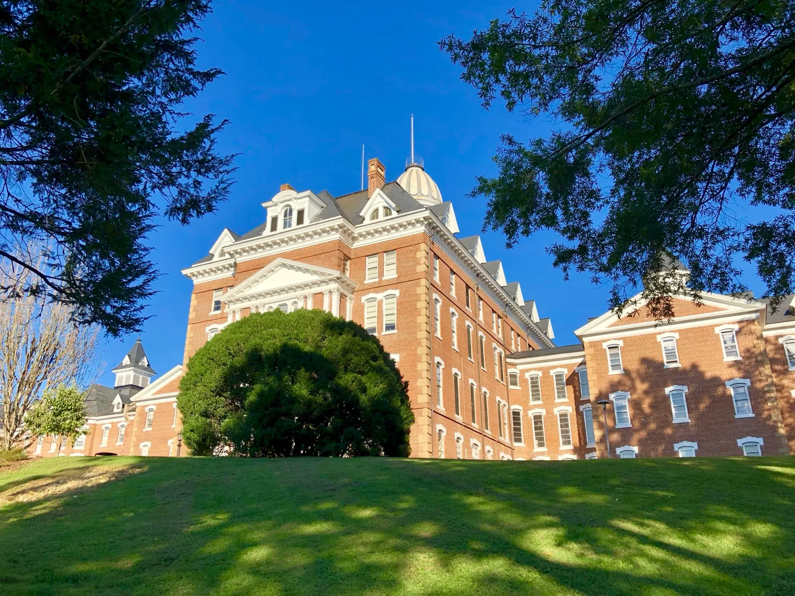 The red brick main building of Broughton Hospital, a state psychiatric hospital in Morganton, North Carolina, behind a round shrub and a sunlit lawn
