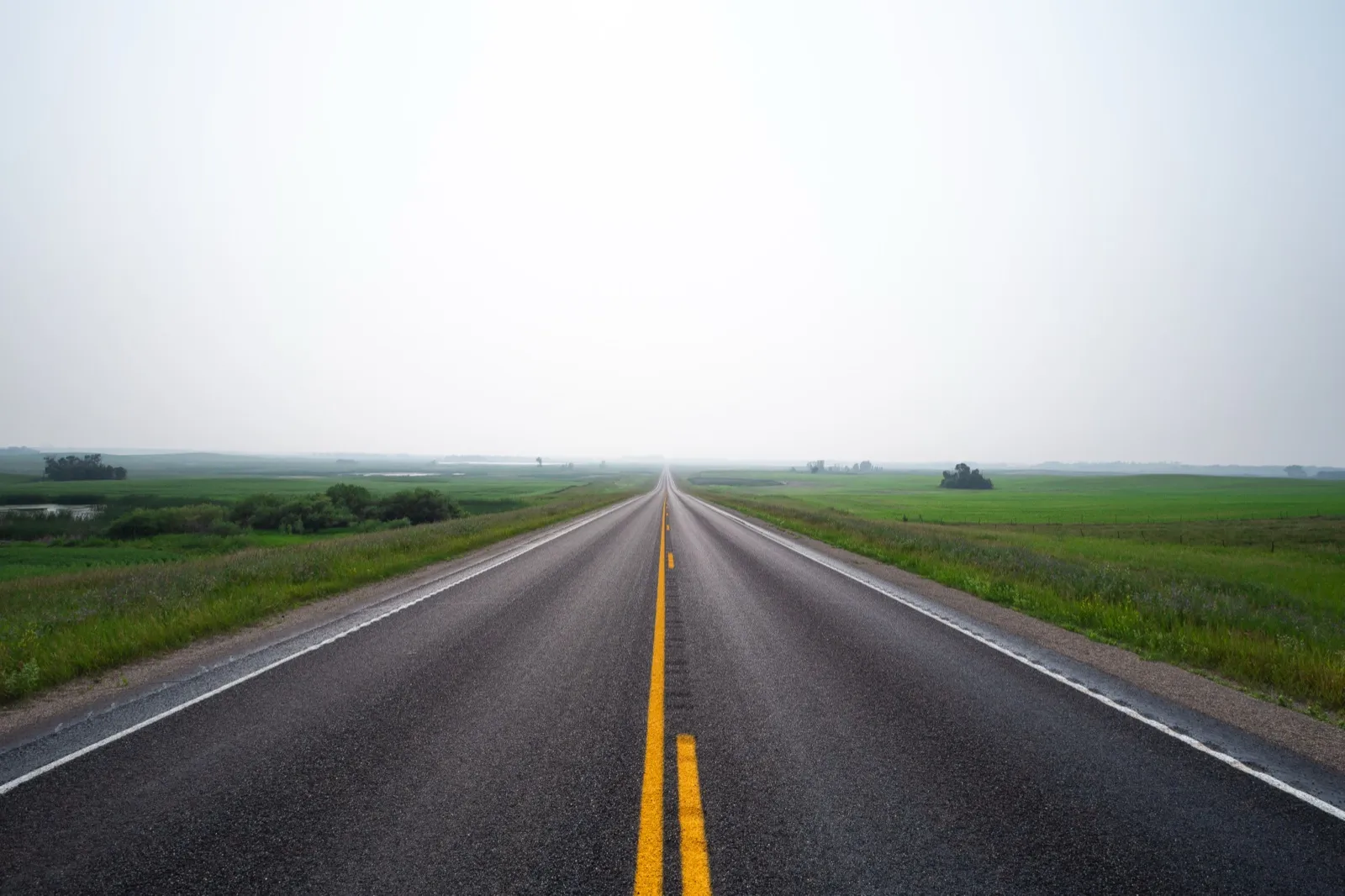 A two-lane highway with yellow center lines running straight across flat green farmland near Wolford, North Dakota
