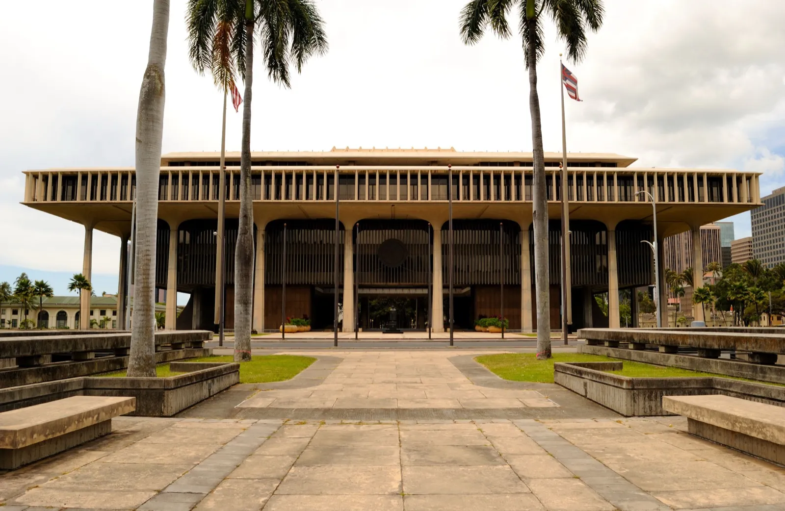 The Hawaii State Capitol in Honolulu seen across a paved plaza, framed by tall palm trees, with flags flying out front