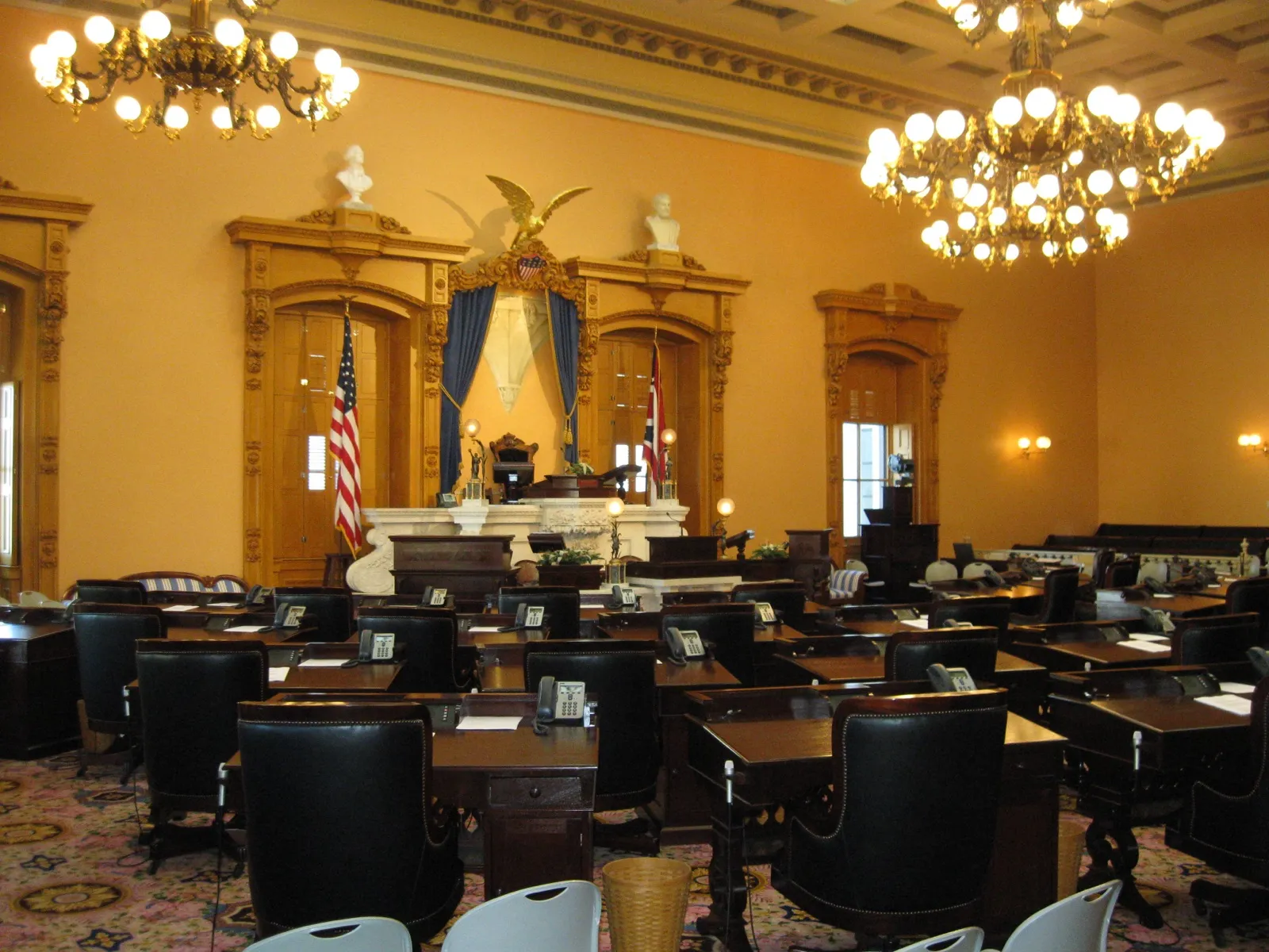 The empty Ohio Senate chamber in the Statehouse, with rows of wooden desks and leather chairs facing the rostrum under two chandeliers