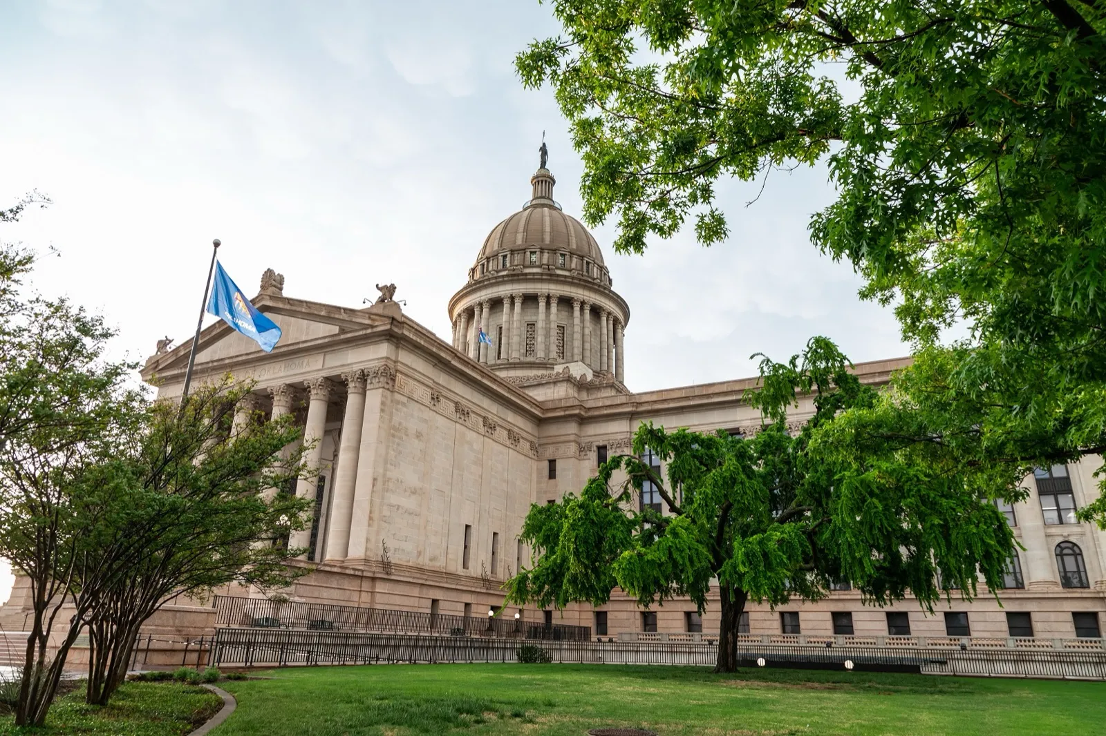 The Oklahoma State Capitol in Oklahoma City, its dome rising behind leafy trees, with the state flag flying on the south lawn