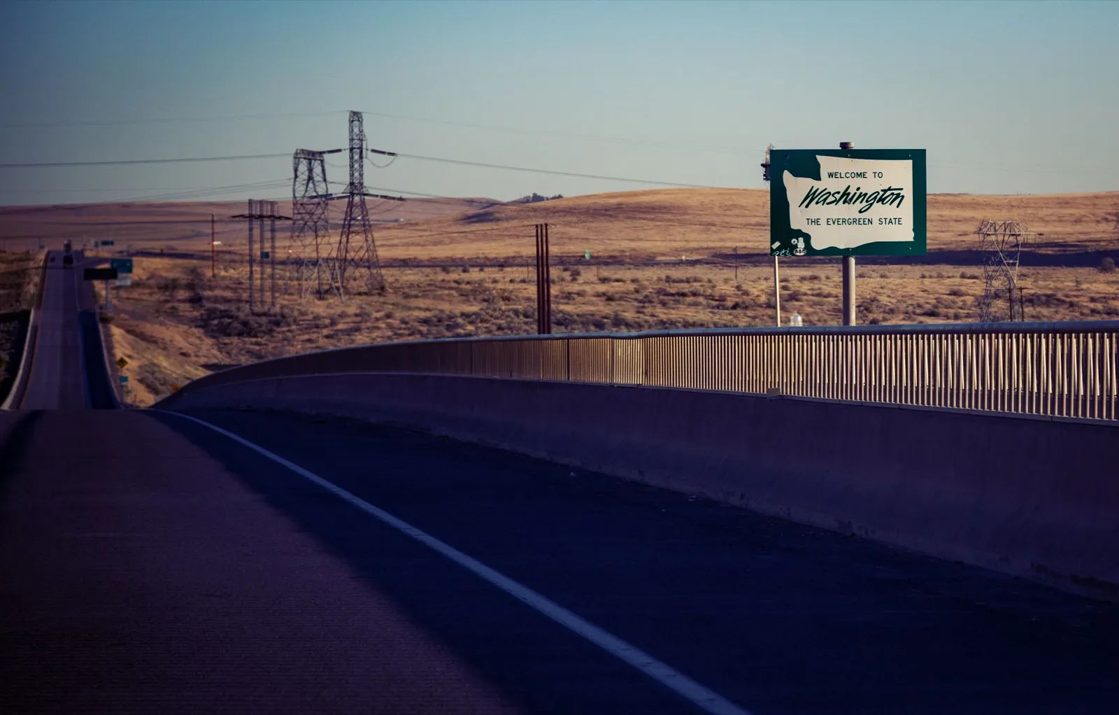 A green Welcome to Washington, The Evergreen State sign beside a highway bridge at the Oregon and Washington state line, with dry hills behind