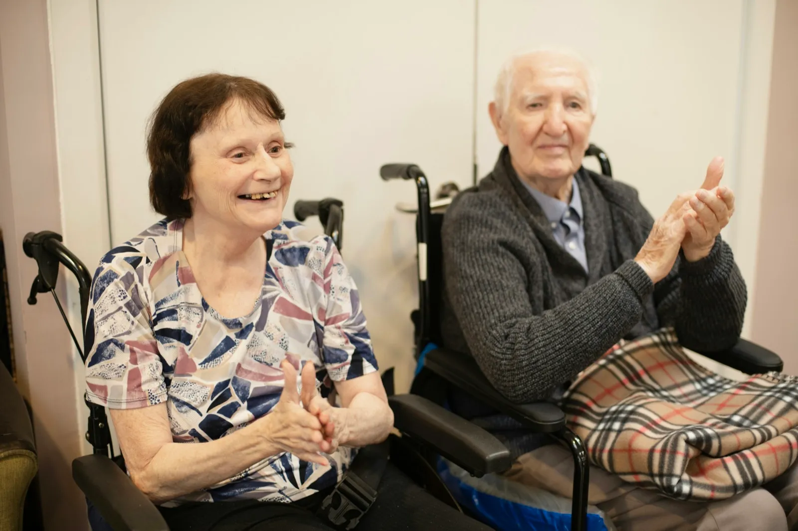Two older adults in wheelchairs smiling and clapping during a group activity in a care center