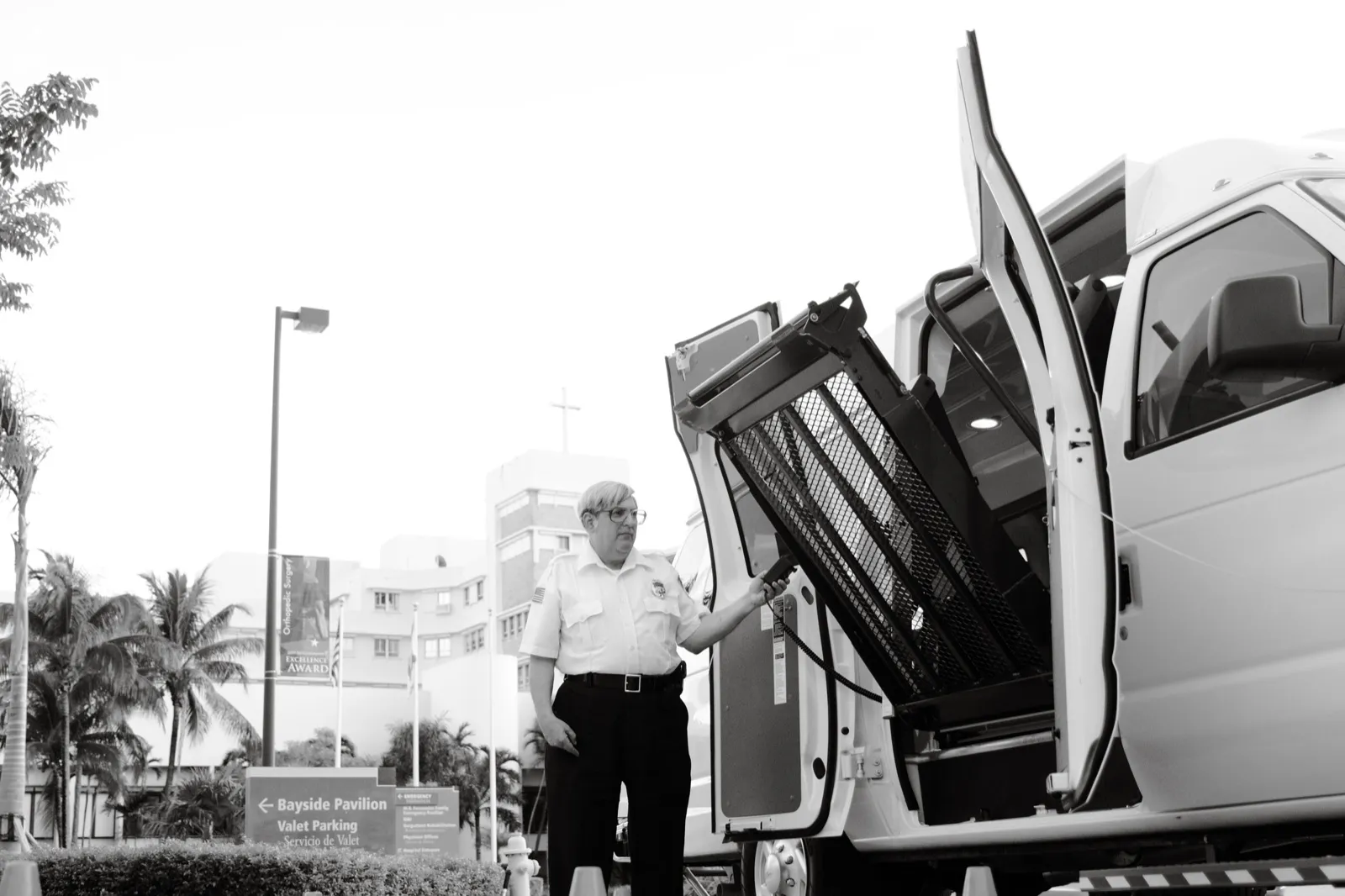 A driver in a white uniform shirt works the wheelchair lift of a white van outside a hospital, in black and white