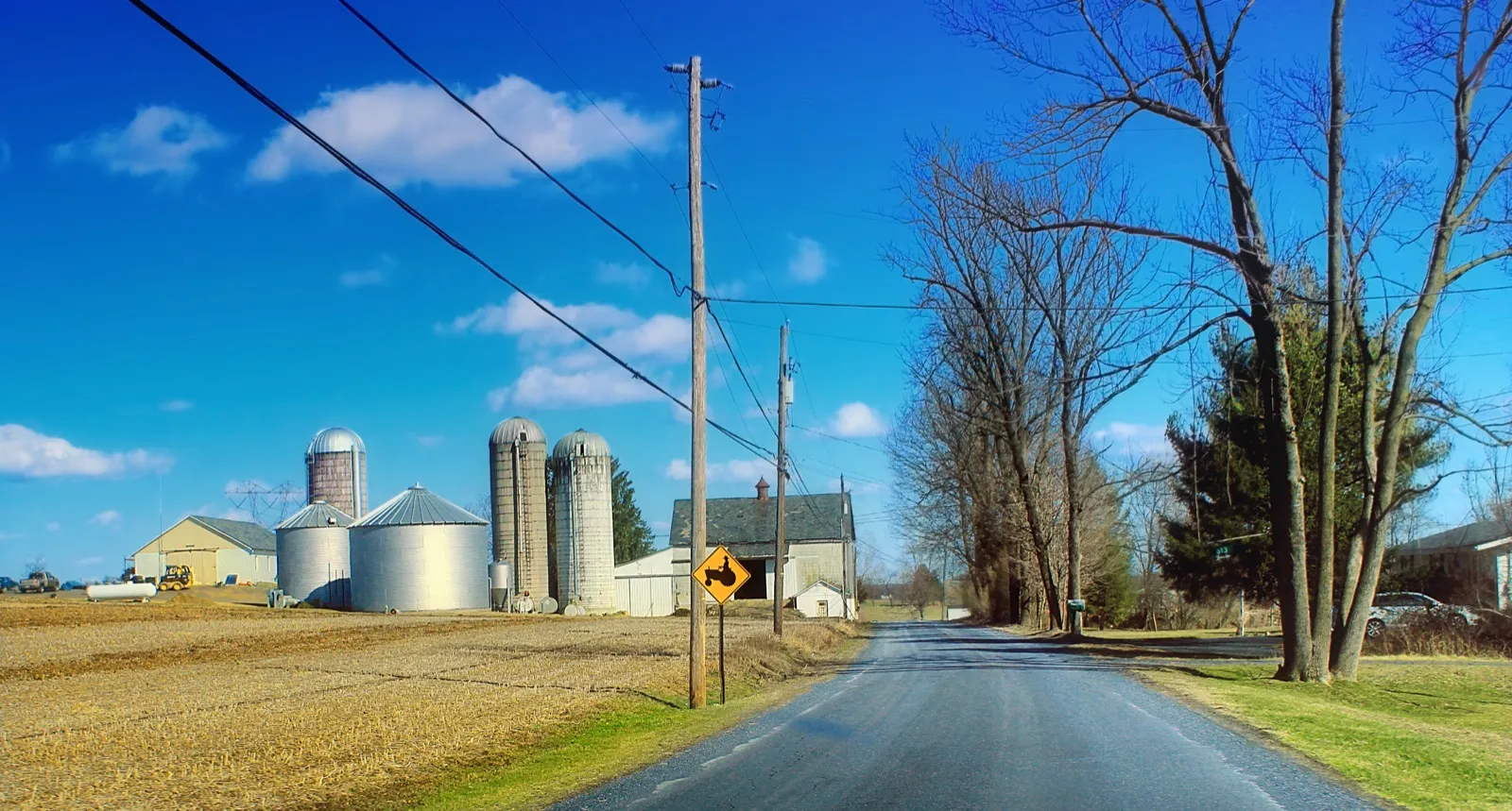 A quiet two-lane country road past a farm with grain silos and a barn in Northampton County, Pennsylvania, under a blue sky