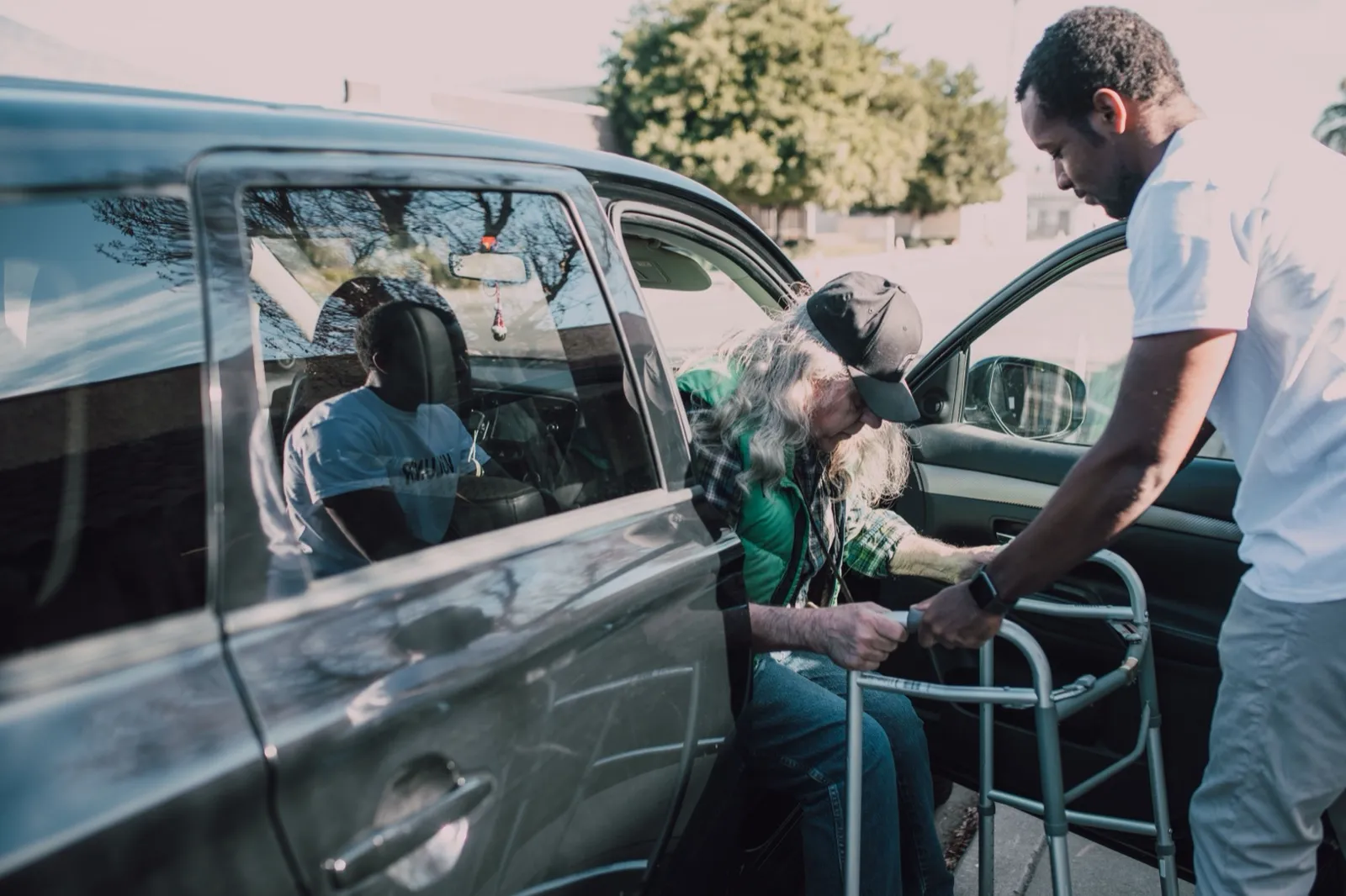 A man holds a folding walker steady at an open car door while an older rider in a cap and green vest gets ready to step out