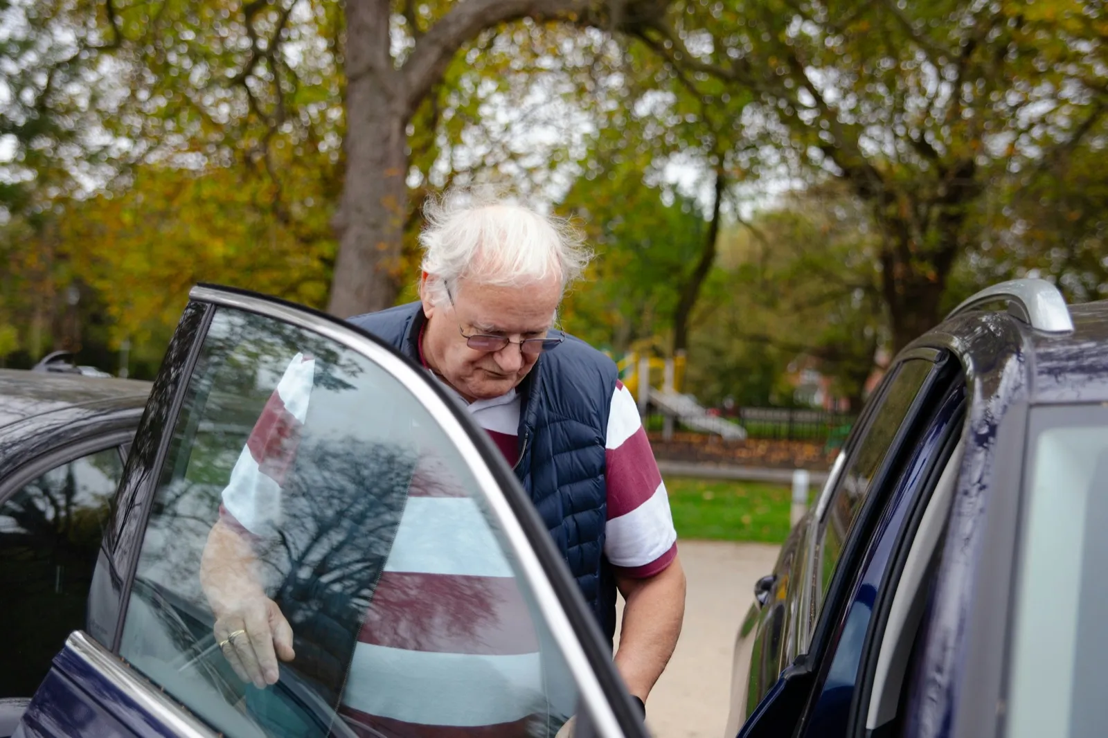 A white-haired man in glasses and a striped shirt opens a car door and steps in, with autumn trees behind him
