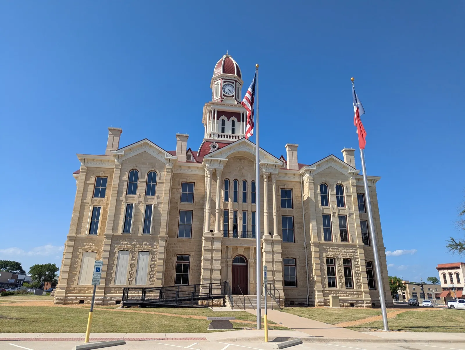 A stone county courthouse with a red-domed clock tower, and United States and Texas flags on tall poles out front under a clear blue sky