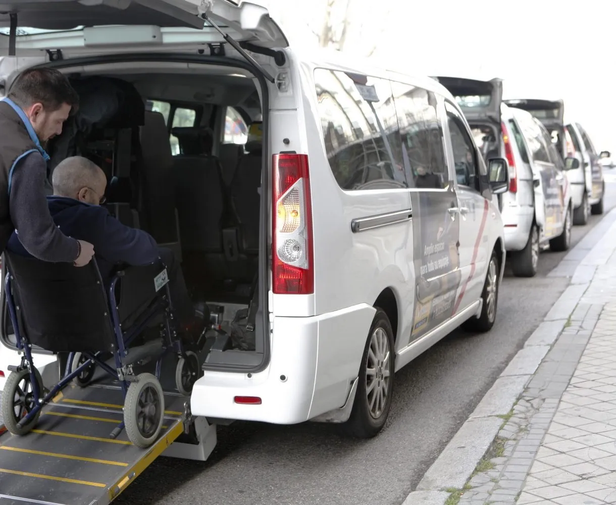 A driver pushes a man in a manual wheelchair up a fold-out ramp into the back of a white van parked at a city curb