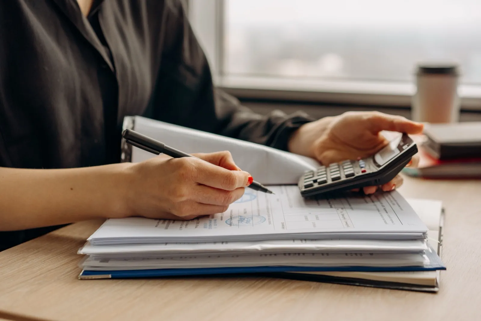 A person at a desk checking a stack of printed statements with a pen in one hand and a calculator in the other