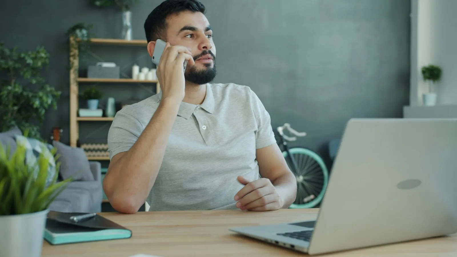A man on a phone call at a table in his living room, with a laptop open in front of him